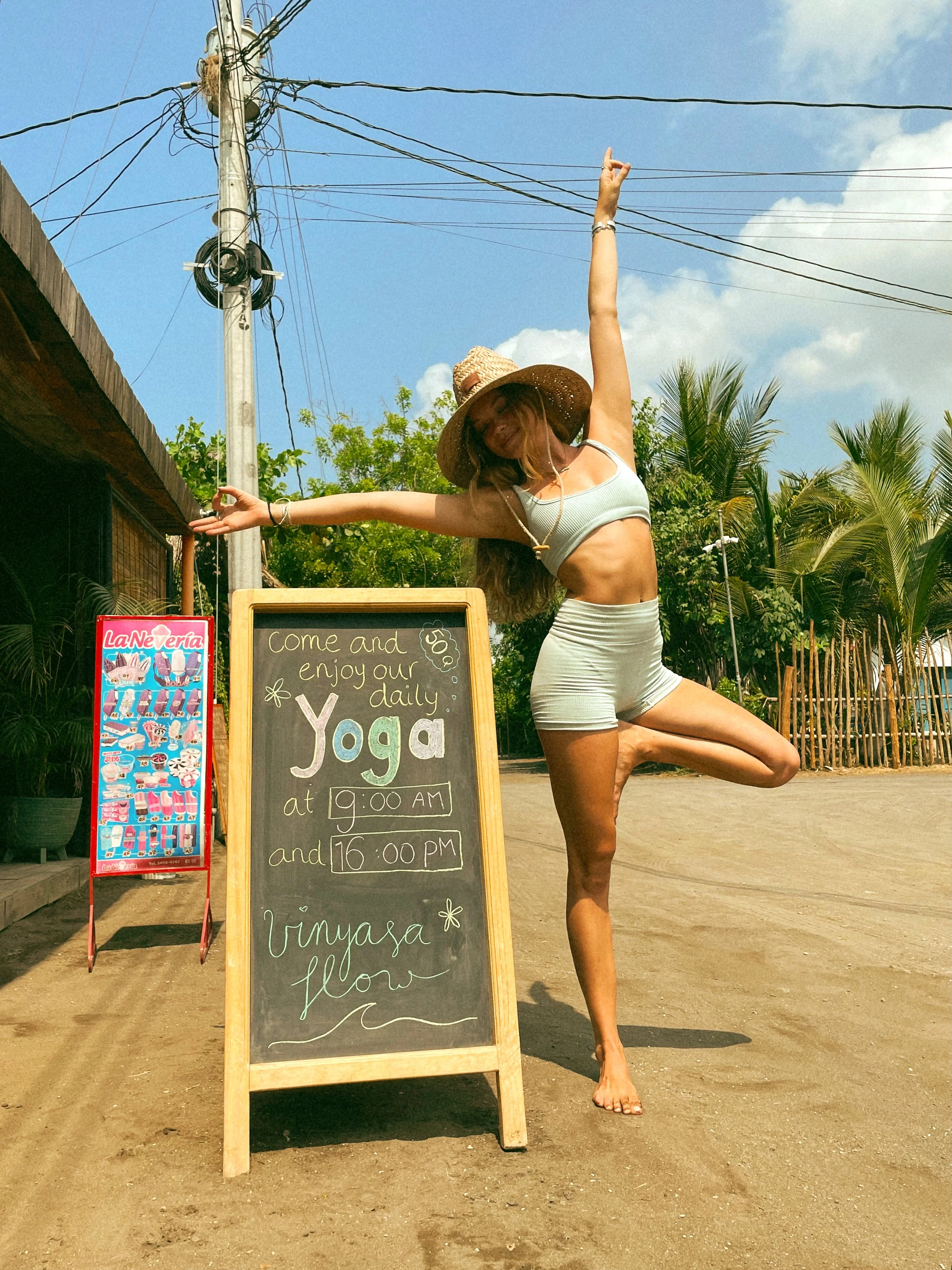 A woman wearing a straw hat and a sleeveless top is doing a yoga pose outdoors next to a chalkboard sign and an ice cream stand, with greenery and electrical wires in the background.