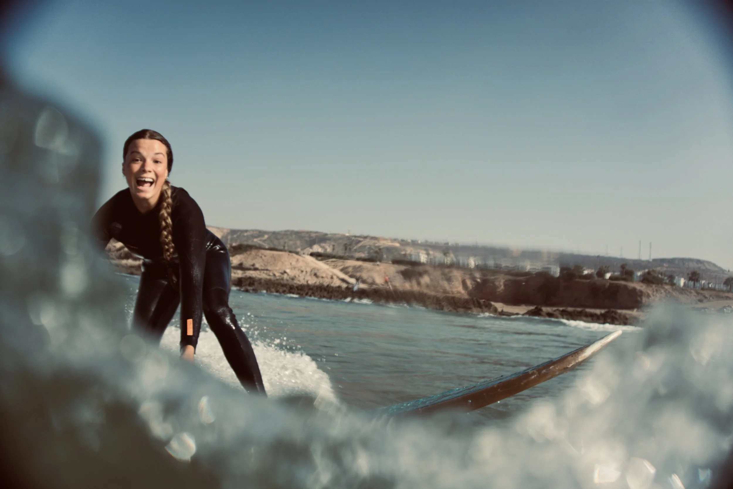 A woman with a braid, smiling, on a surfboard near the shoreline with rocky cliffs and blue sky in the background.