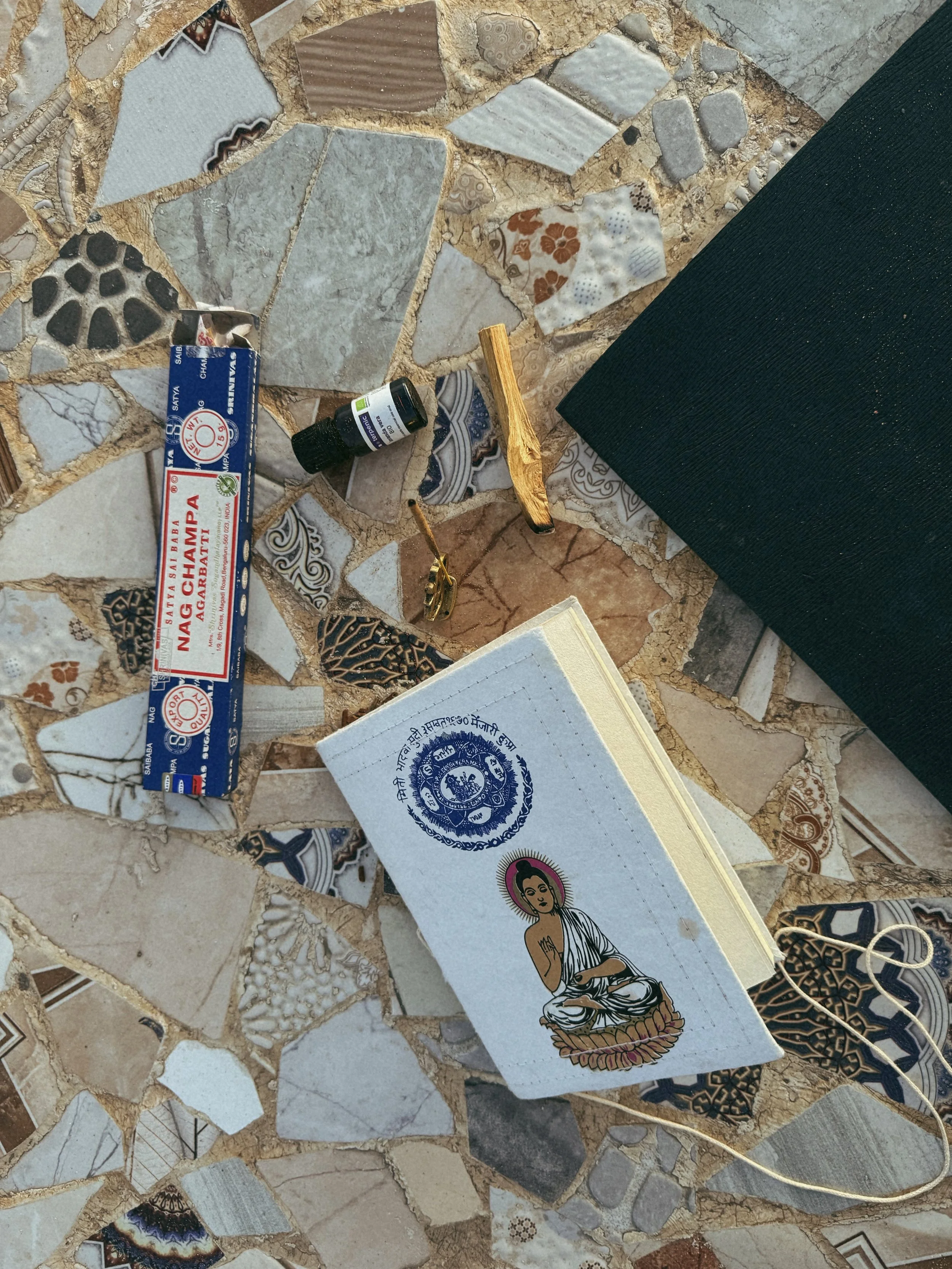 Various objects on a mosaic tiled surface, including a box of incense sticks, a small container, a gold-colored fork, a notebook with a Buddha illustration, and prayer flags.
