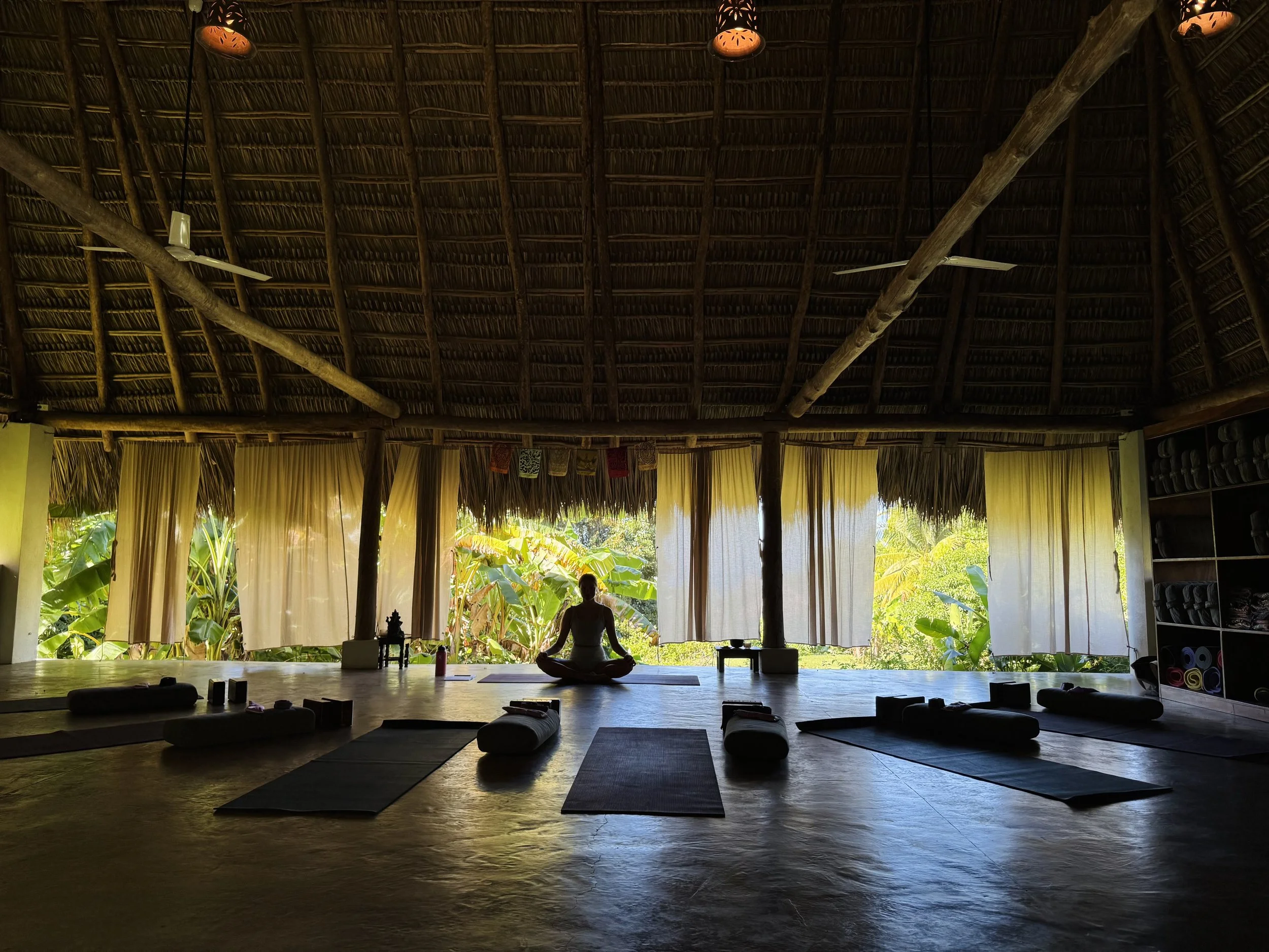 Interior of a rustic open-air yoga studio with a thatched roof, curtains, and lush greenery outside. A person is sitting cross-legged in a meditation pose on a yoga mat in the center.