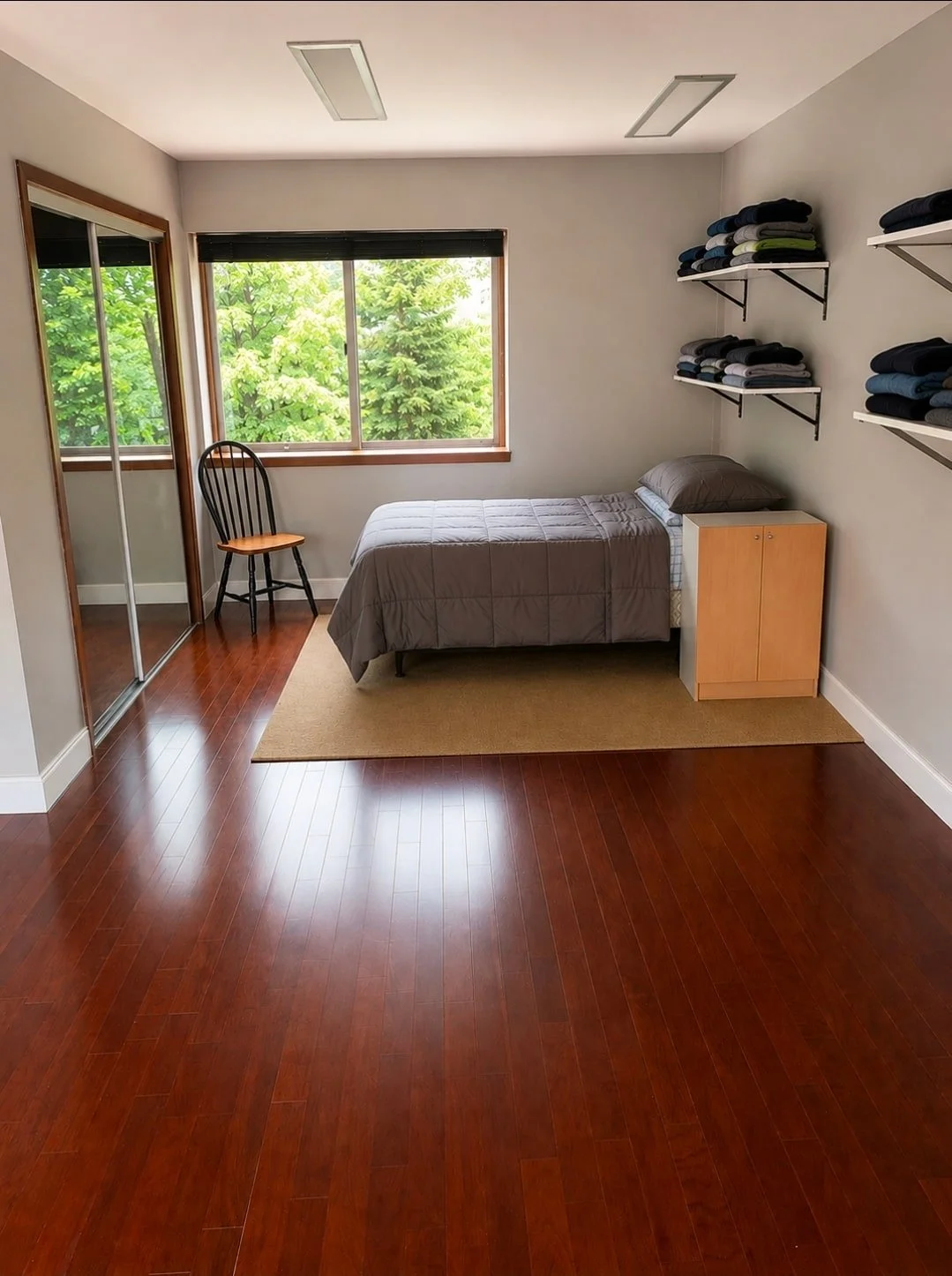 A room at Crosswalk Recovery House featuring a bed, a mirrored, sliding closet door, shelves, a chair, a nightstand, and hardwood floors.