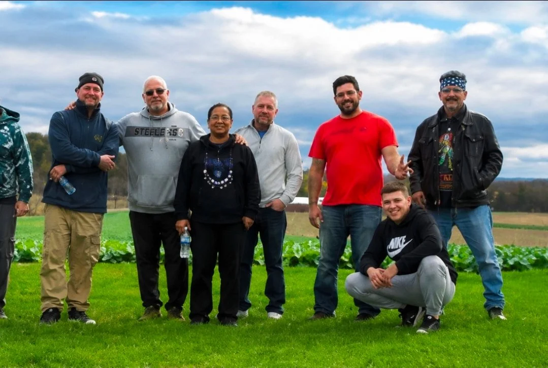Group of eight men, residents of Crosswalk Recovery House, standing on a grassy field with farmland and cloudy sky in the background.