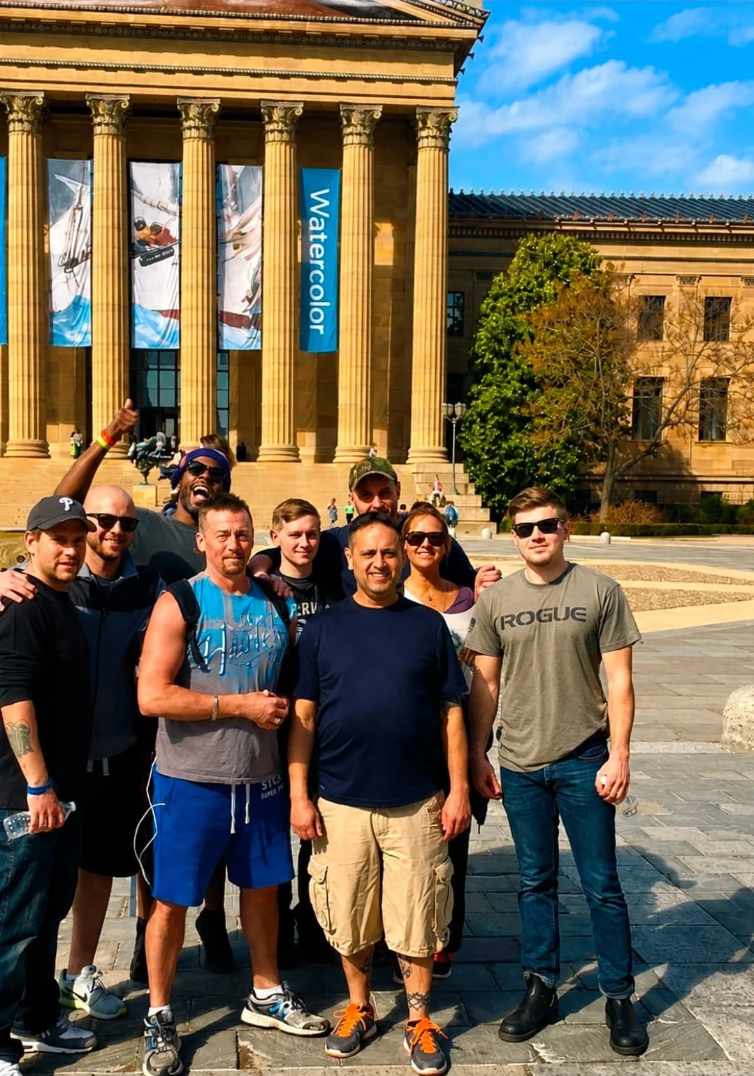 Group of men, residents of Crosswalk Recovery House on a day trip, standing in front of the Philadelphia Museum of Art, with tall columns, blue banners, and a sign that says 'Watercolor', outdoors on a sunny day.