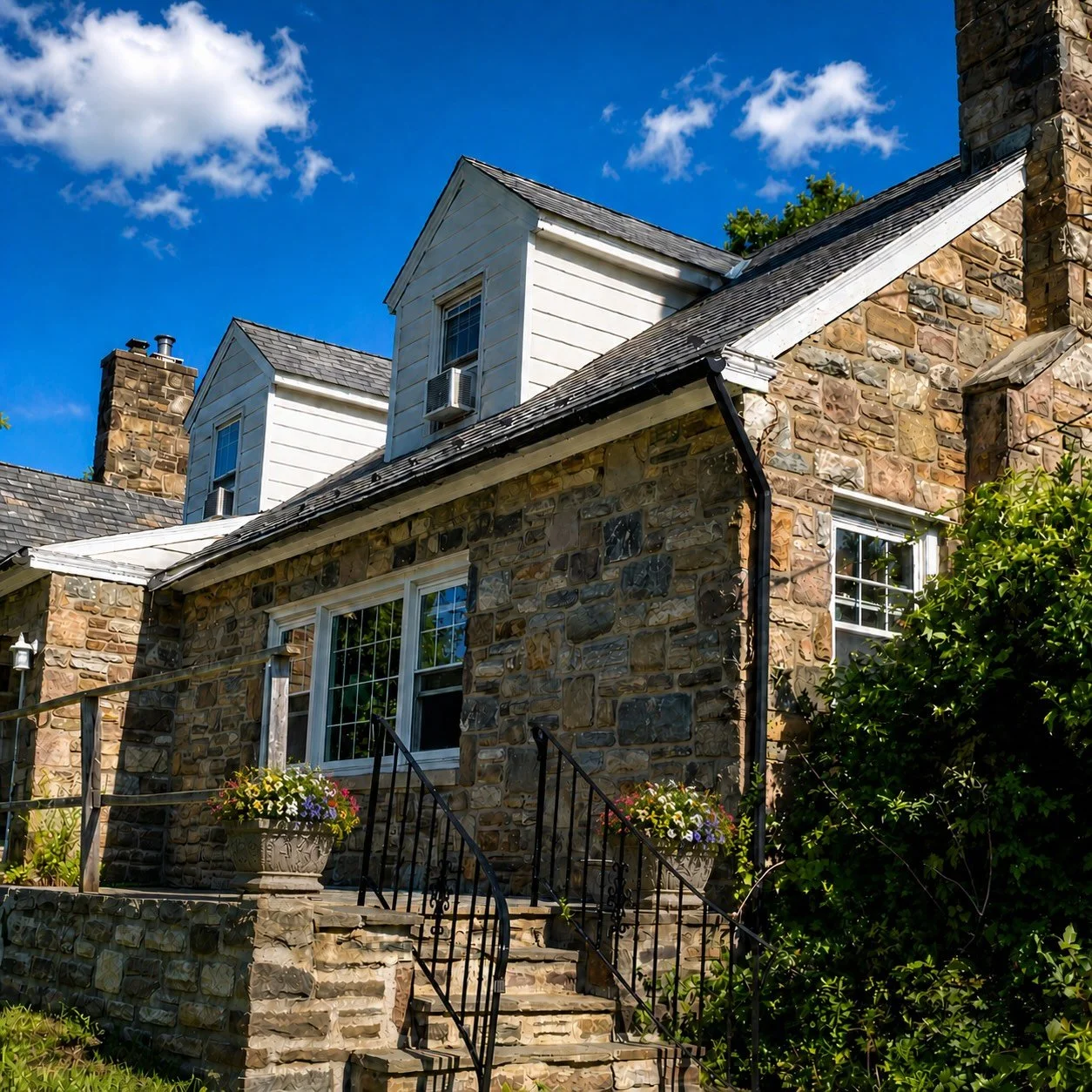 The Crosswalk Recovery House in Bucks County, Pennsylvania, is a stone house and features white dormer windows showing stairs with black handrails leading up to a front door with planters on either side filled with colorful flowers.