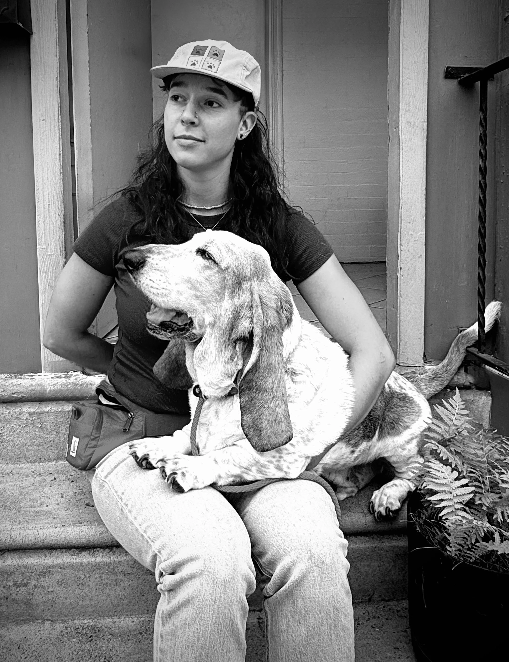 A young woman with long dark hair, wearing a baseball cap with paw prints, a black t-shirt, and light-colored pants, sitting outdoors with a large Labrador Retriever dog, both appearing relaxed.