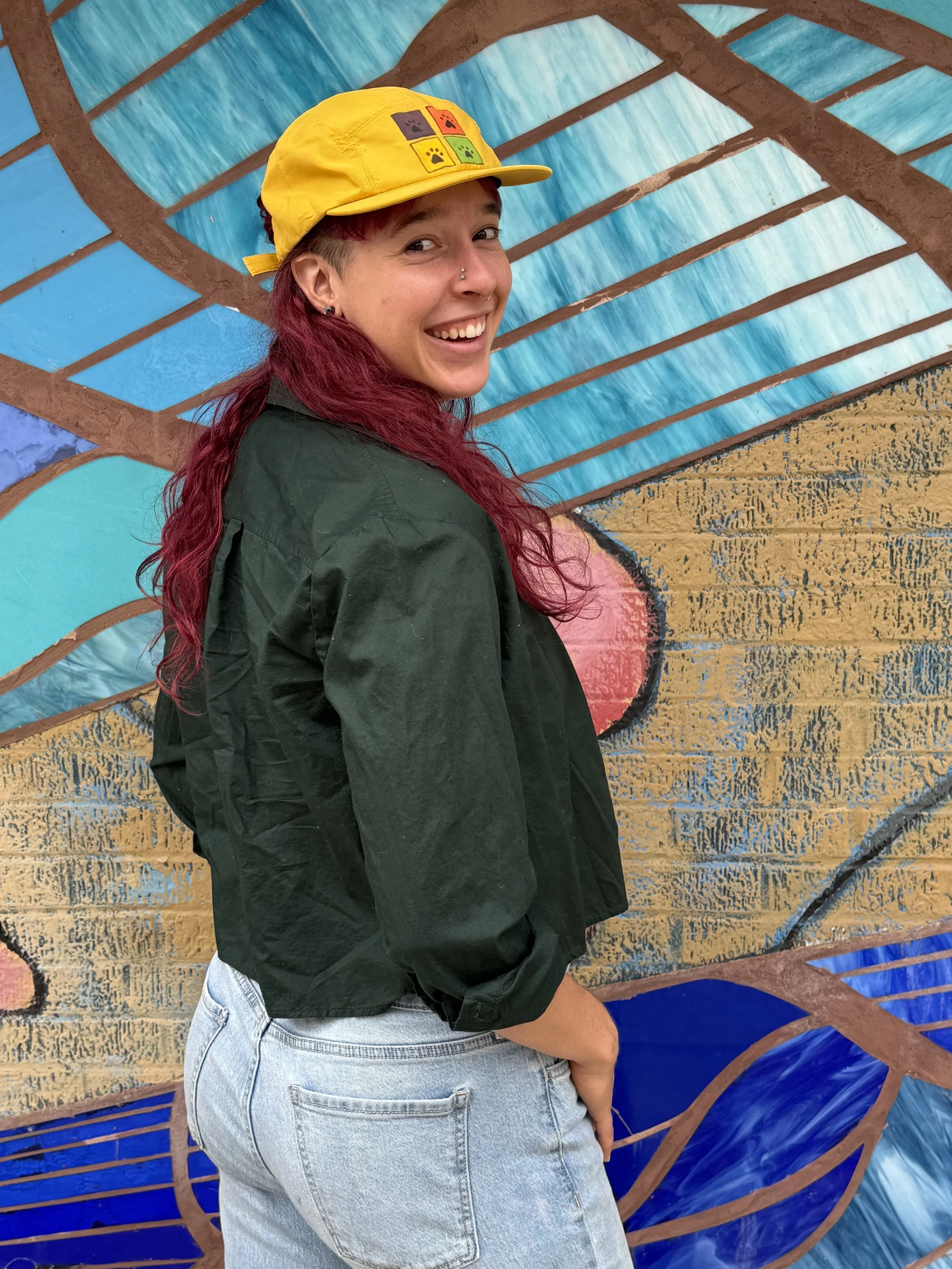 A woman with long red hair wearing a yellow cap, black jacket, and gray jeans standing in front of a colorful mural with blue, brown, pink, and yellow sections and geometric patterns.