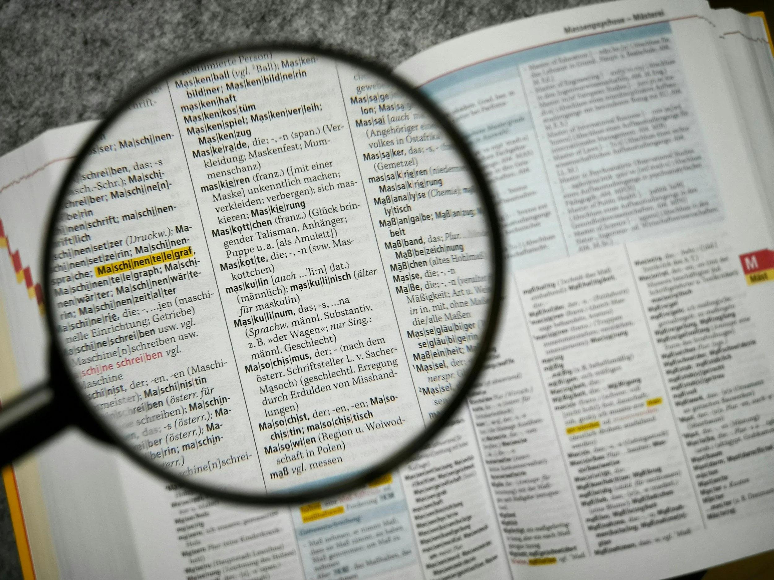 A close-up view of a dictionary page focused through a magnifying glass, highlighting the German words 'Maschinenlehrgraf' and 'Maschinenlehrtable.' The magnifying glass enlarges these words, while the surrounding text remains clear.