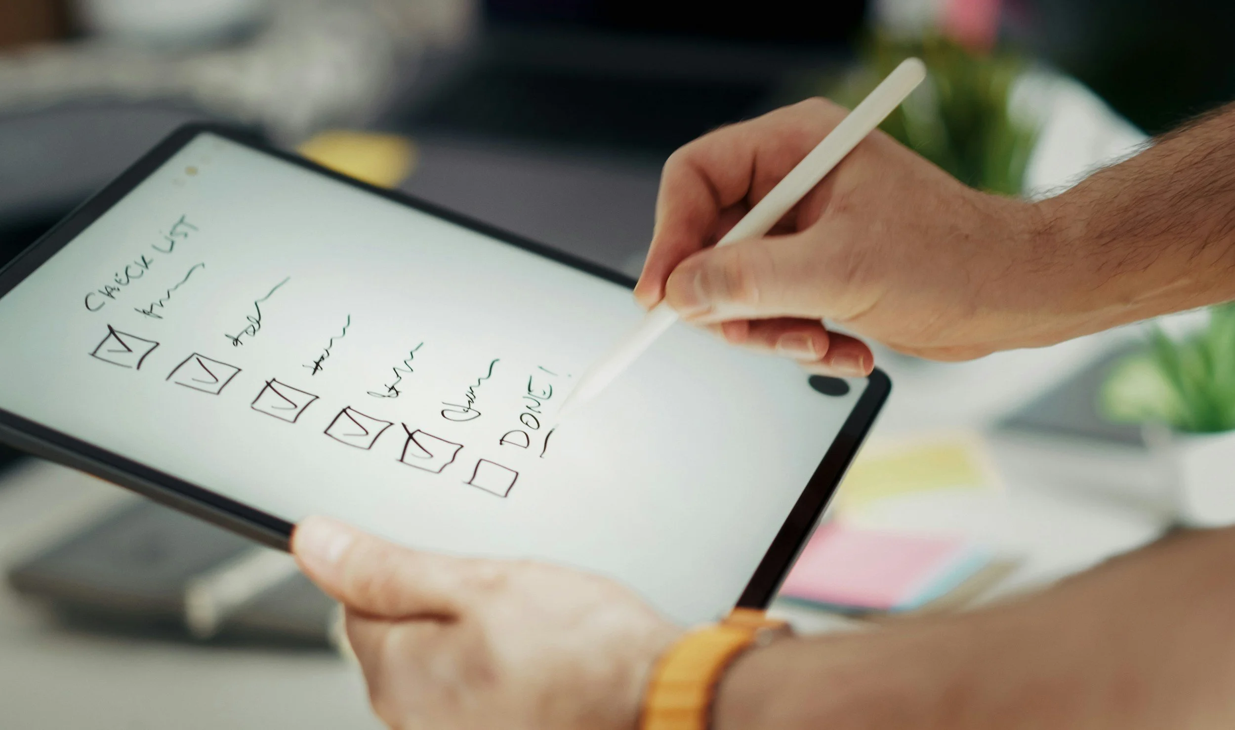 A person holding a digital tablet with a handwritten to-do list that includes checkboxes, titled 'Checklist'. The hand is using a stylus to interact with the screen, and the person is wearing a wristwatch on their left wrist. The background is slightly blurred with hints of greenery.