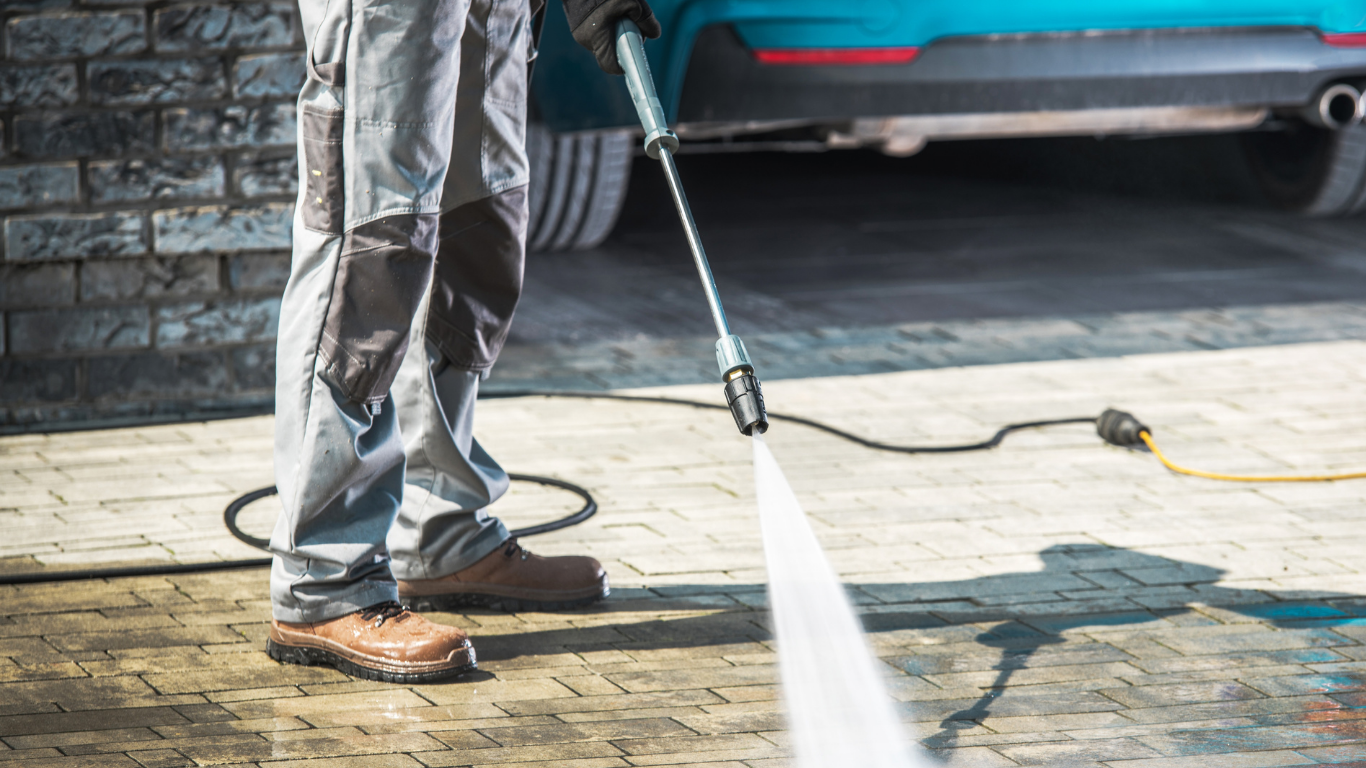 Person wearing work boots and gray work pants pressure washing a paved brick driveway with a high-pressure hose.