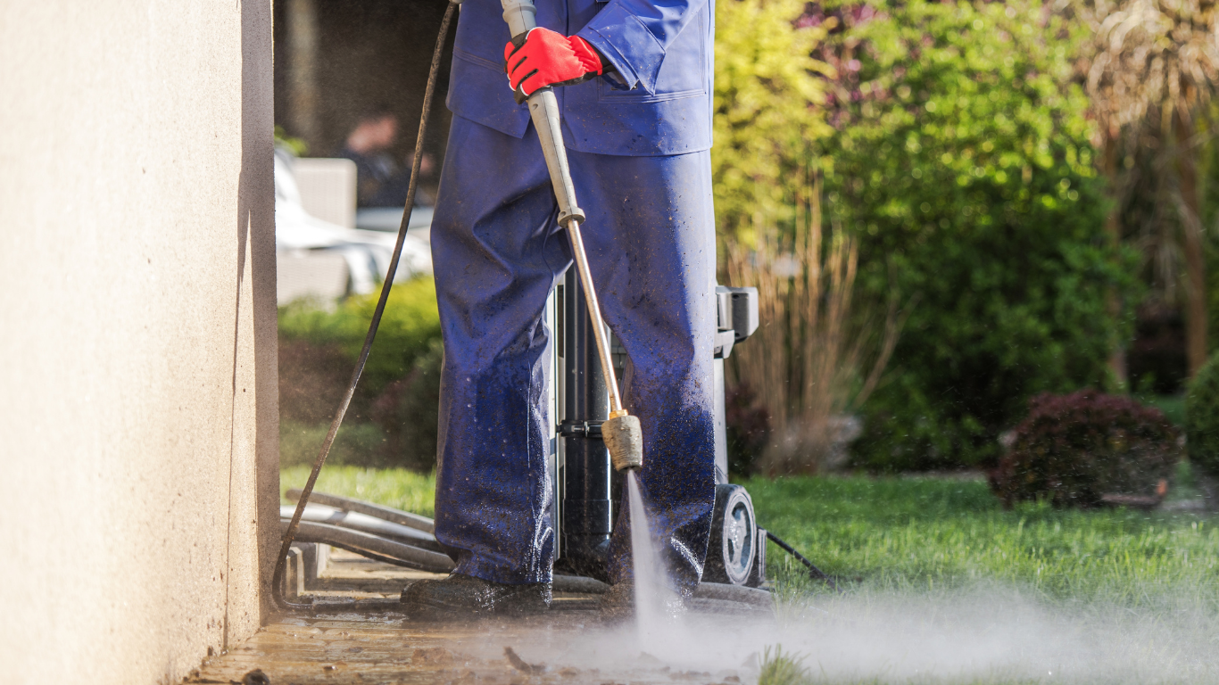 A person wearing blue overalls and red gloves power washing a concrete surface next to a beige wall in a garden.