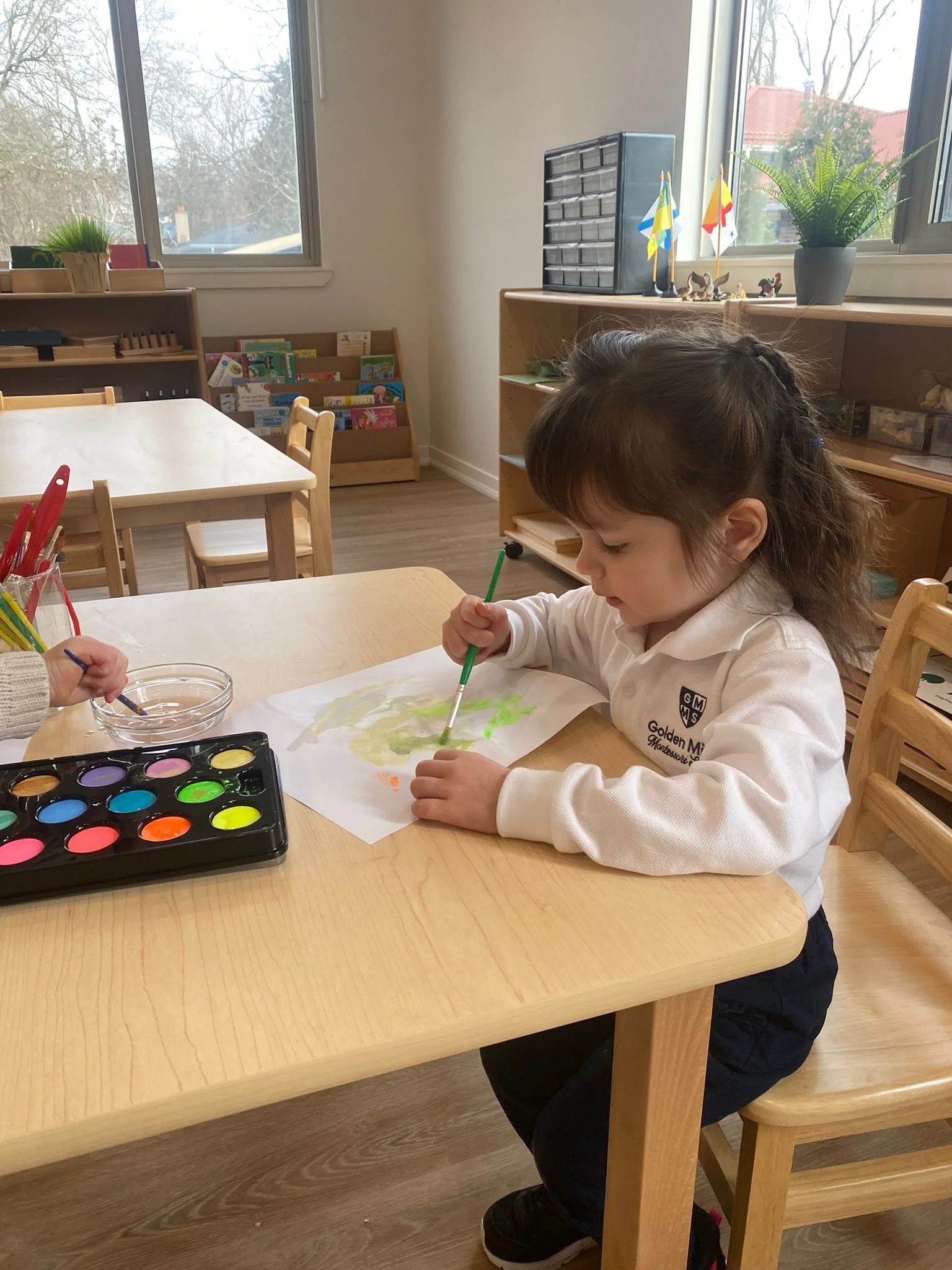 A young girl sitting at a wooden table, painting on a sheet of paper with watercolors. In the background, there are windows, a bookshelf with books, and small flags and figurines on a shelf.