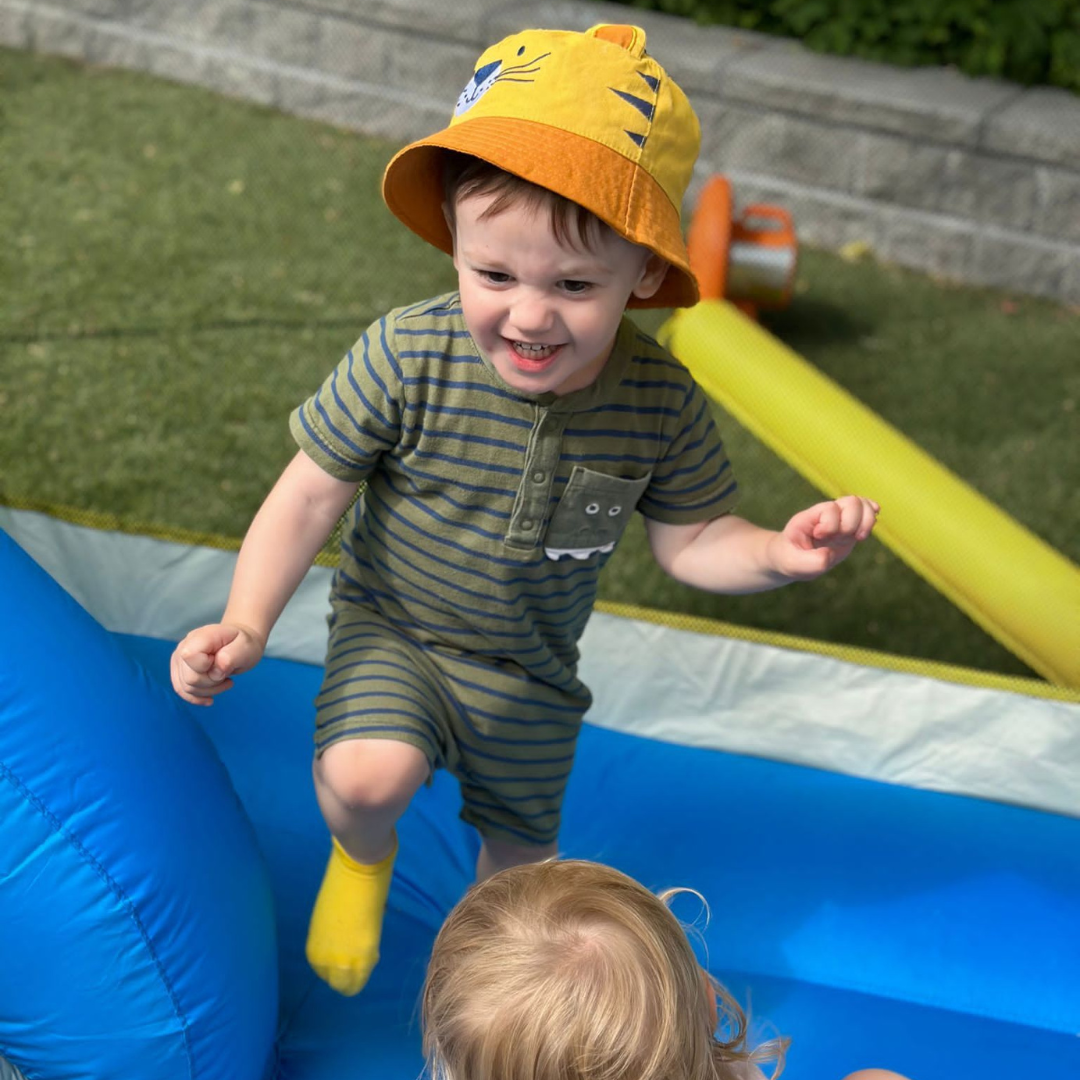 A young boy in a striped shirt and yellow rain boots playing in an inflatable bounce house with a girl inside, wearing a colorful hat.