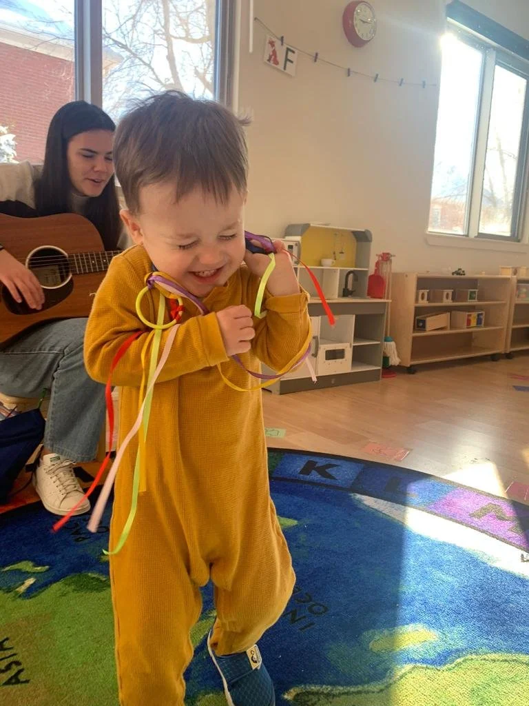A young boy in a yellow outfit is crying and holding colorful streamers, while a woman in the background plays guitar and sings in a classroom with a rug, shelves, and windows.