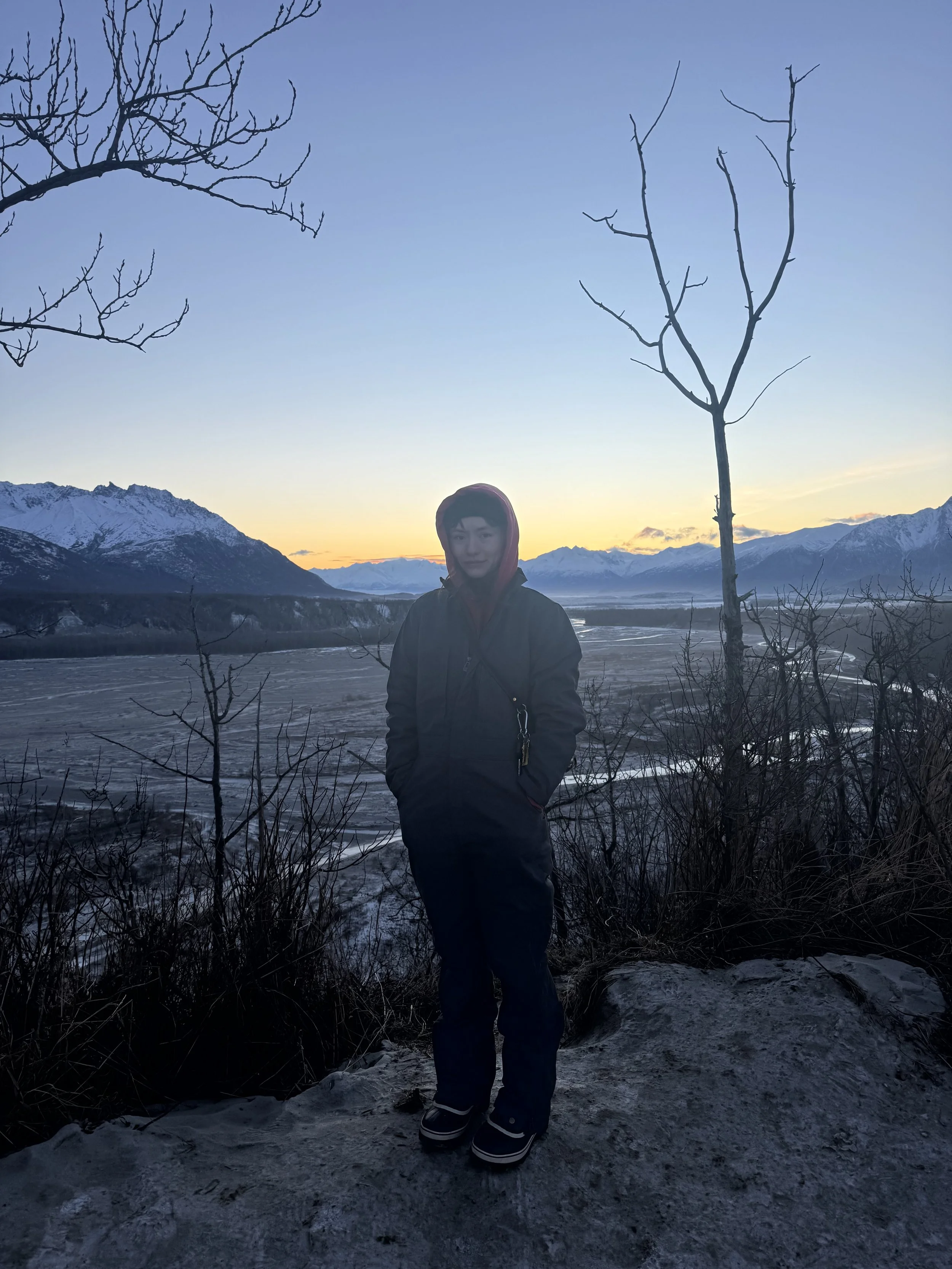 A person standing outdoors on a snowy, rocky terrain with a mountain range and a river in the background during sunset. The person is dressed in winter clothing, including a hooded jacket and snow boots, with leafless trees around.