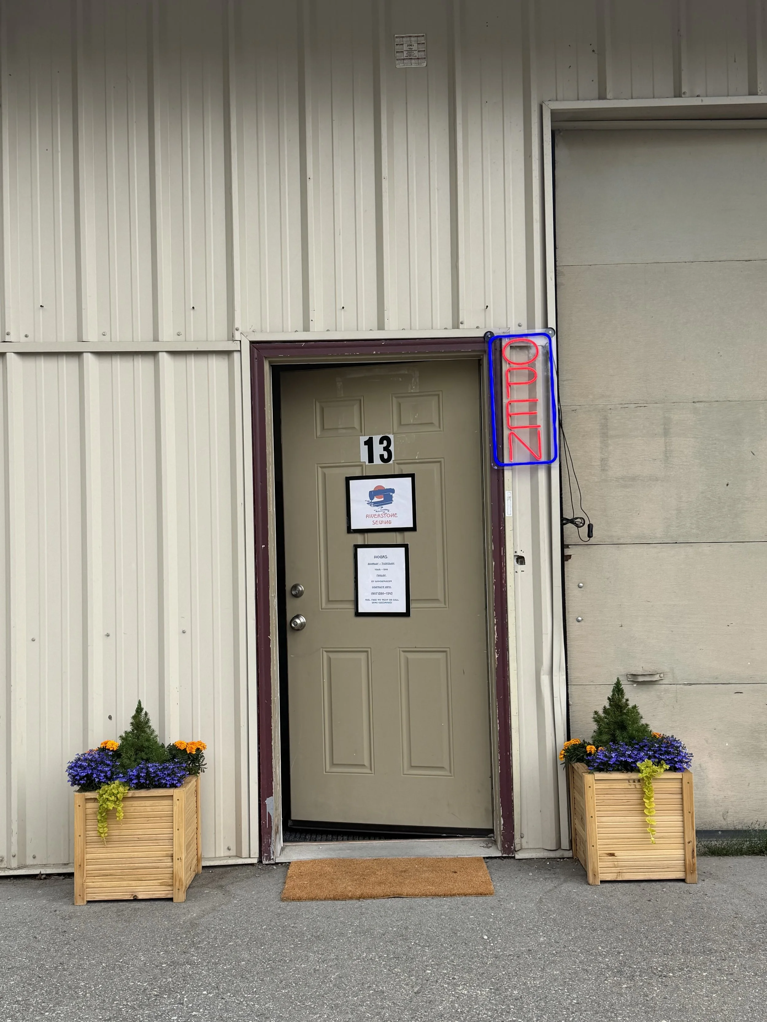 A beige door with a sign that says 'Riverside Sewing,' flanked by two planters with purple and yellow flowers, a doormat in front, and a neon sign that reads 'OPEN' attached to the wall.