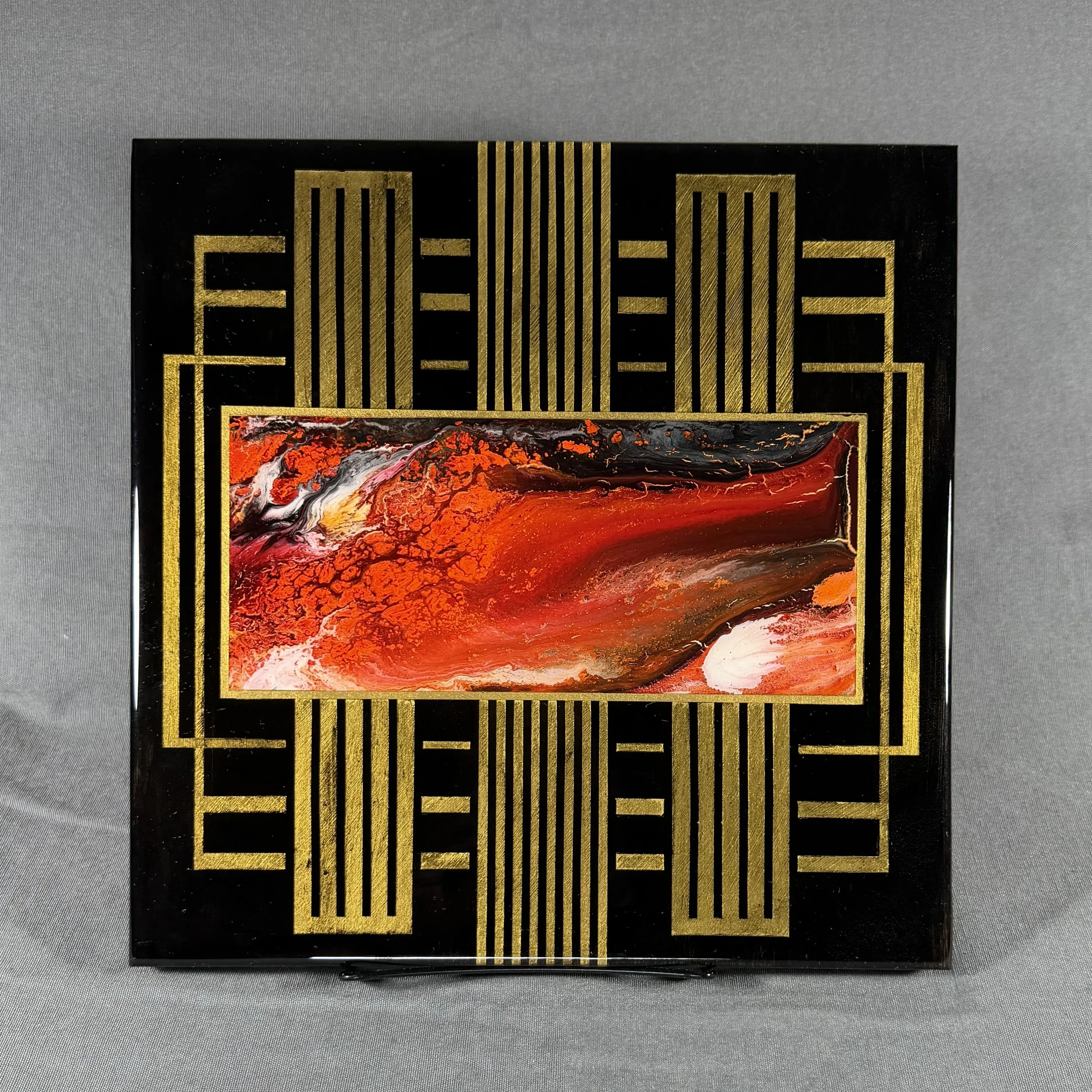 A rectangular decorative piece with a black background and gold geometric pattern framing a central abstract painting with red, orange, white, and black swirls and textures.