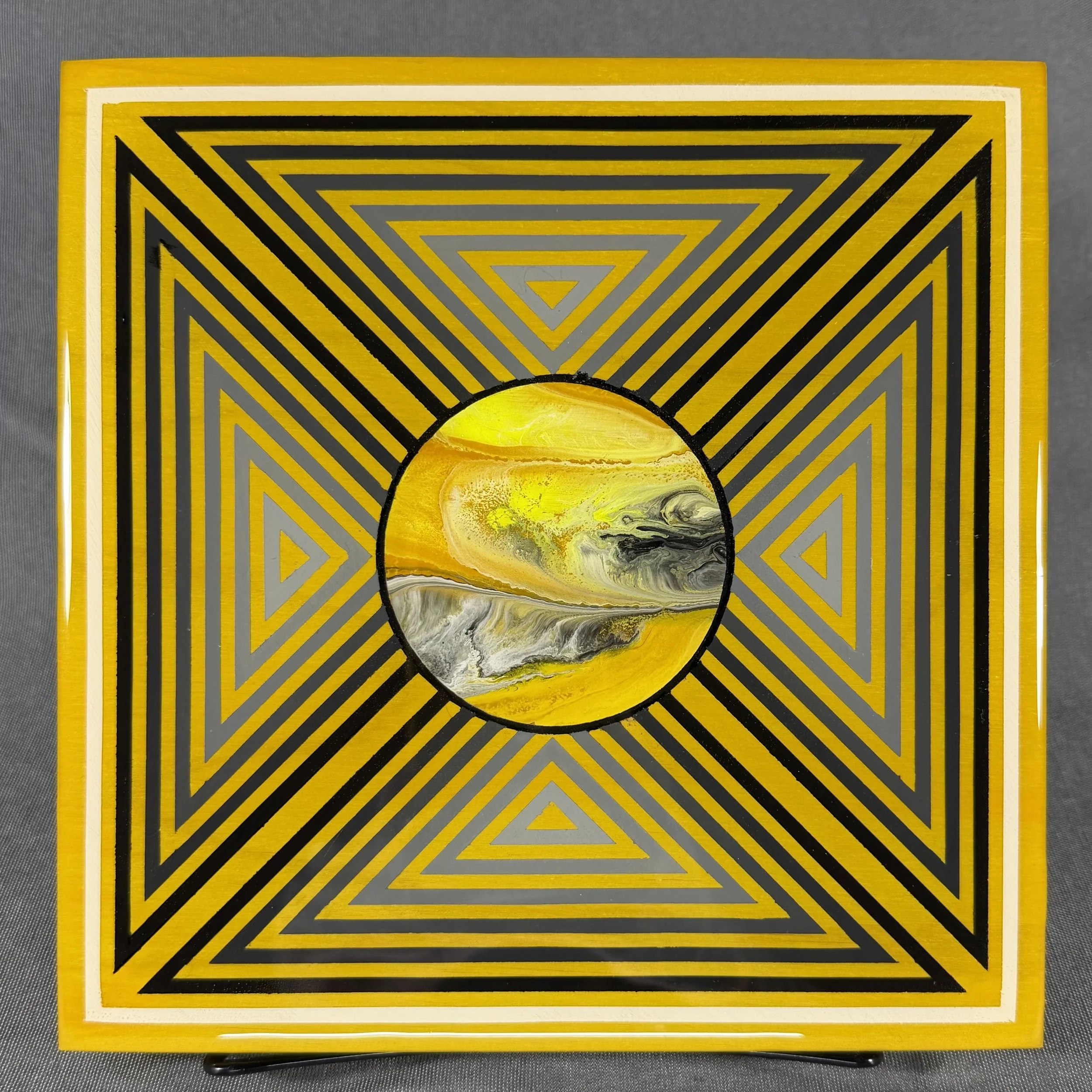 A rectangular artwork with a yellow border featuring black and grey concentric triangles and a central sphere with yellow, black, and grey marbled patterns.