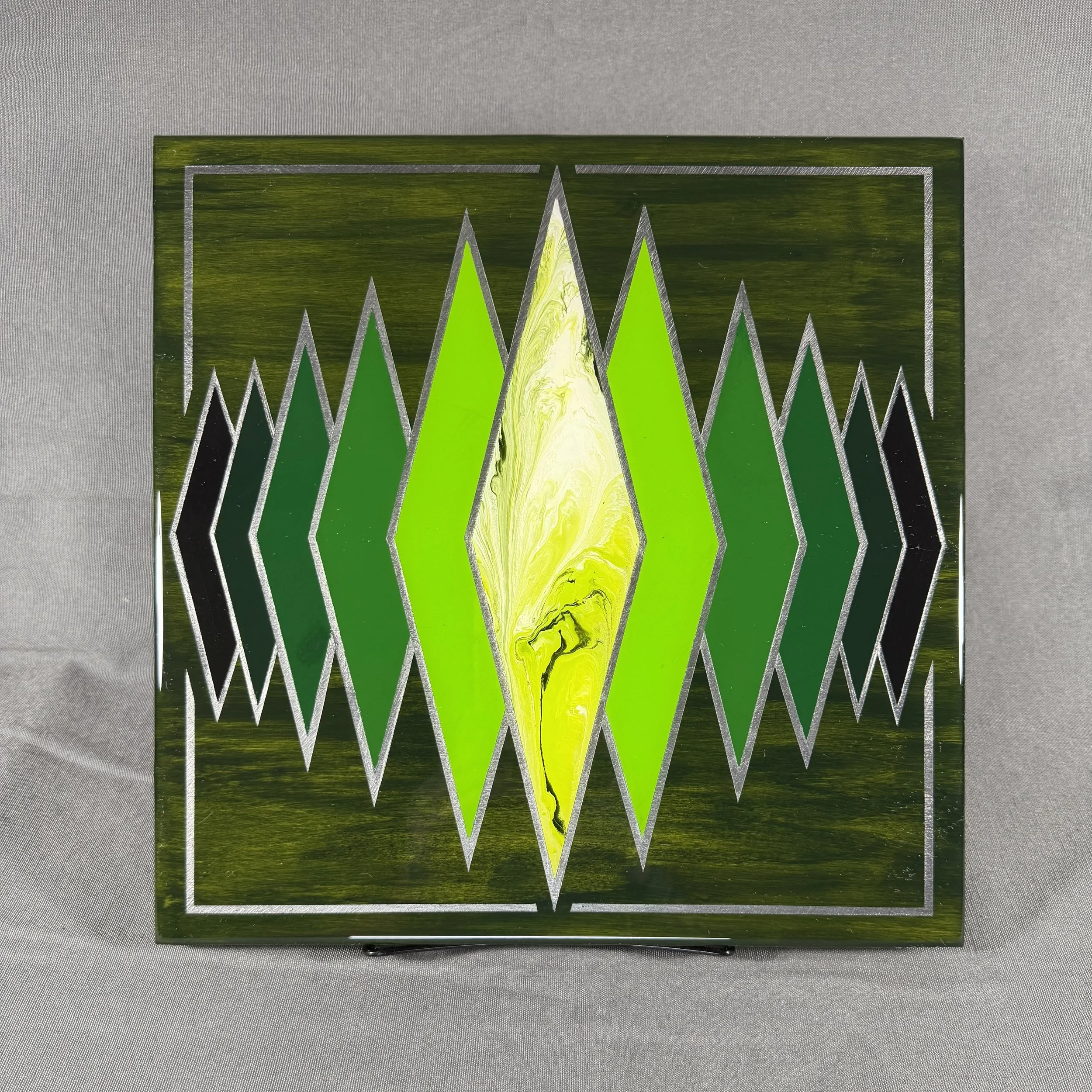 a decorative glass block with a geometric pattern of sharp triangles in neon green, yellow, and black, outlined by silver lines, placed on a gray surface