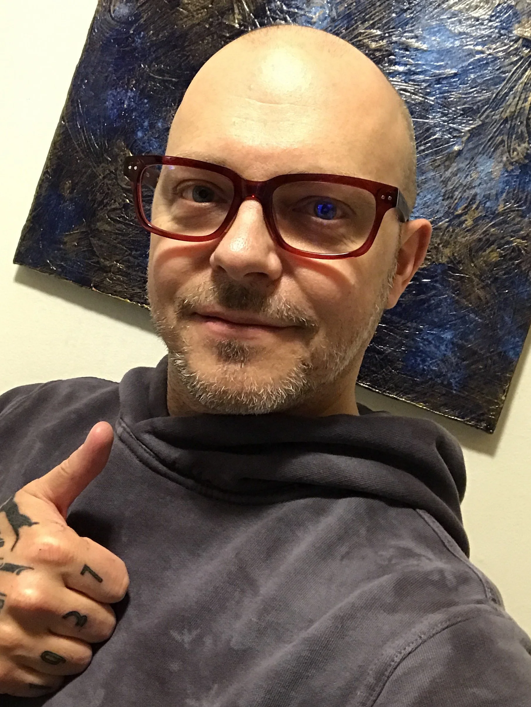 A man with glasses and facial hair taking a selfie, pointing to his tattooed fingers, with a textured dark art piece in the background.