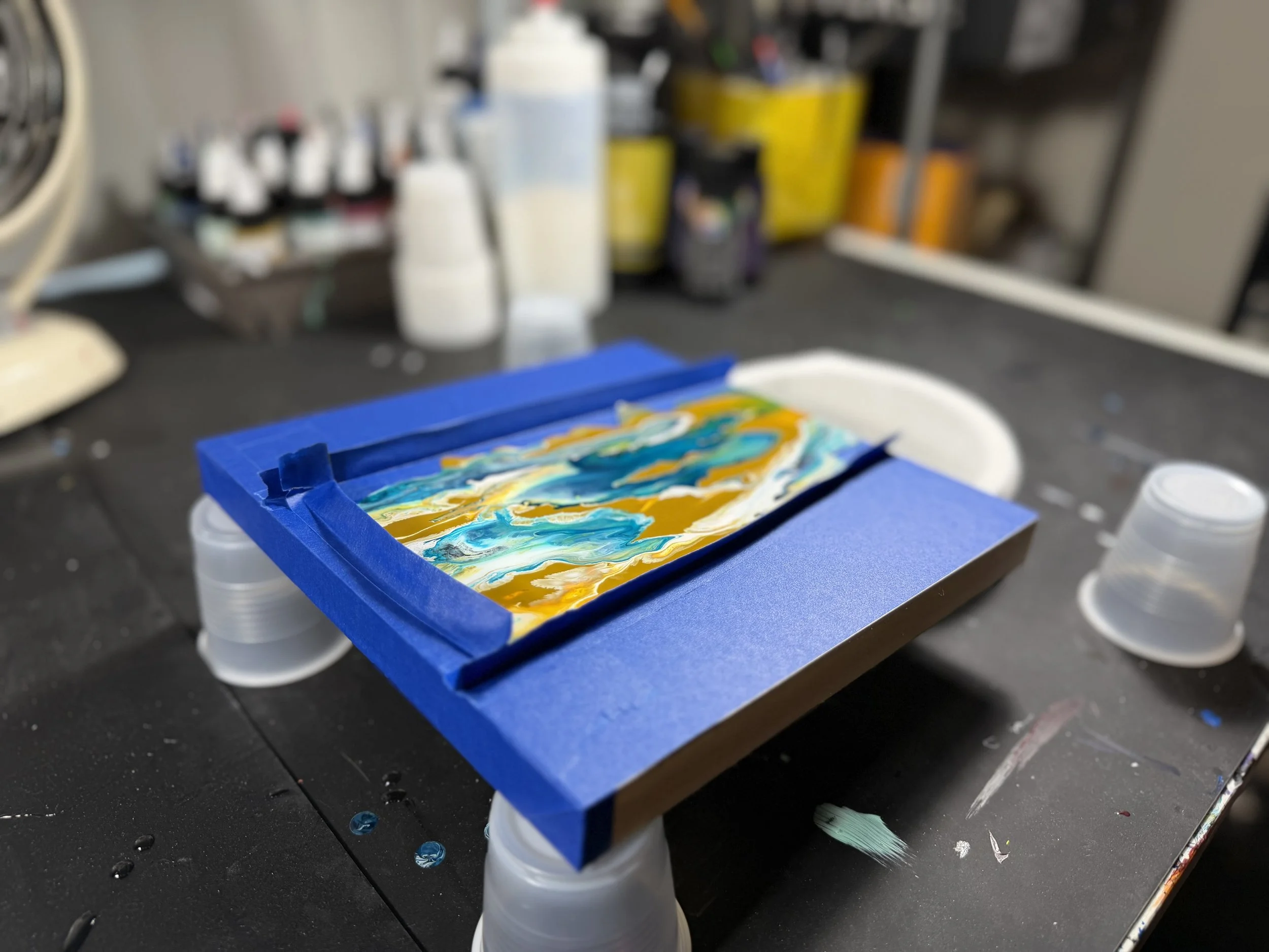 A piece of artwork with blue, yellow, orange, green, and white swirls, placed on a blue frame, on a black table with art supplies in the background.