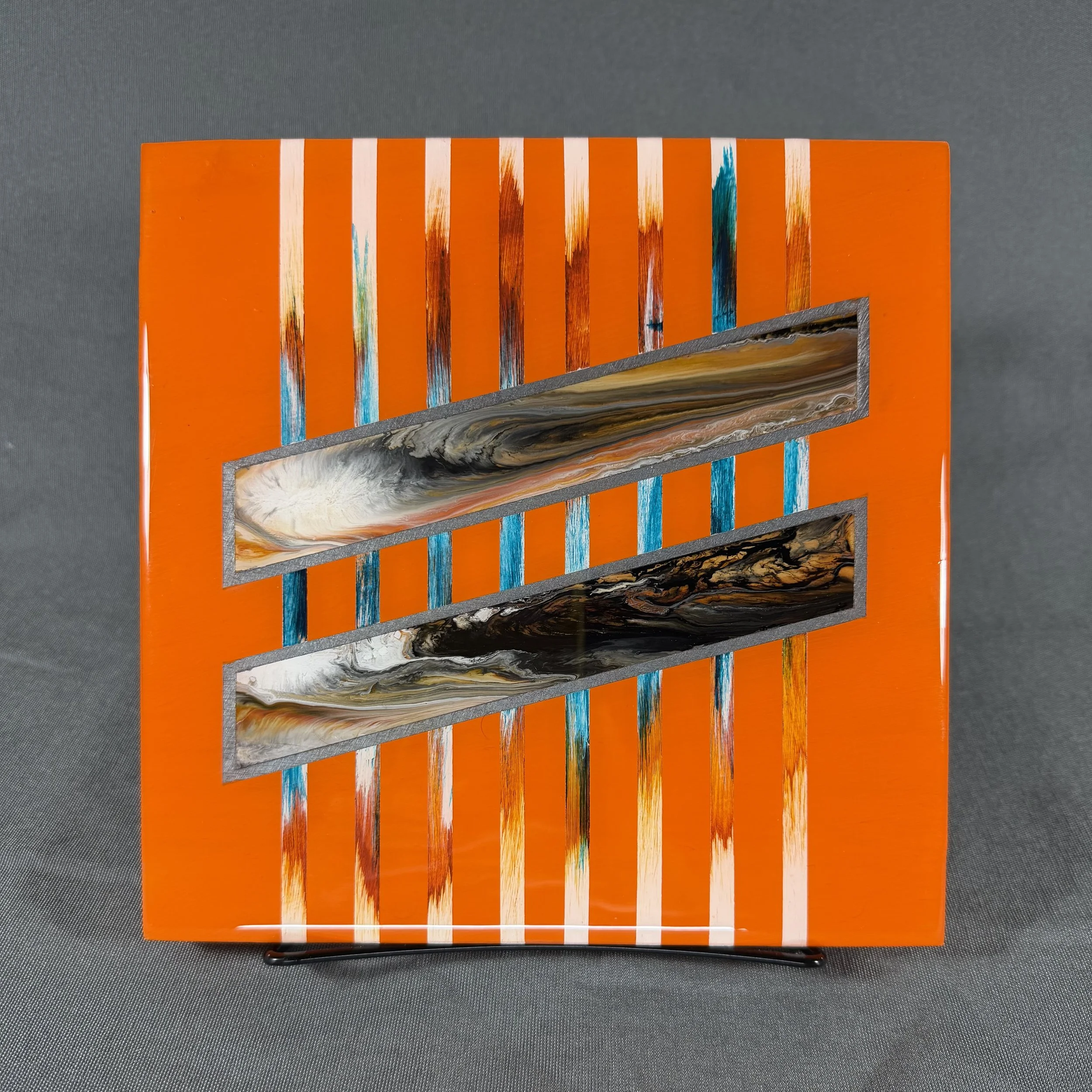 Close-up of a colorful abstract art piece on a framed canvas, featuring bright orange background with vertical streaks of white, blue, and brown paint, leaning against a gray surface.