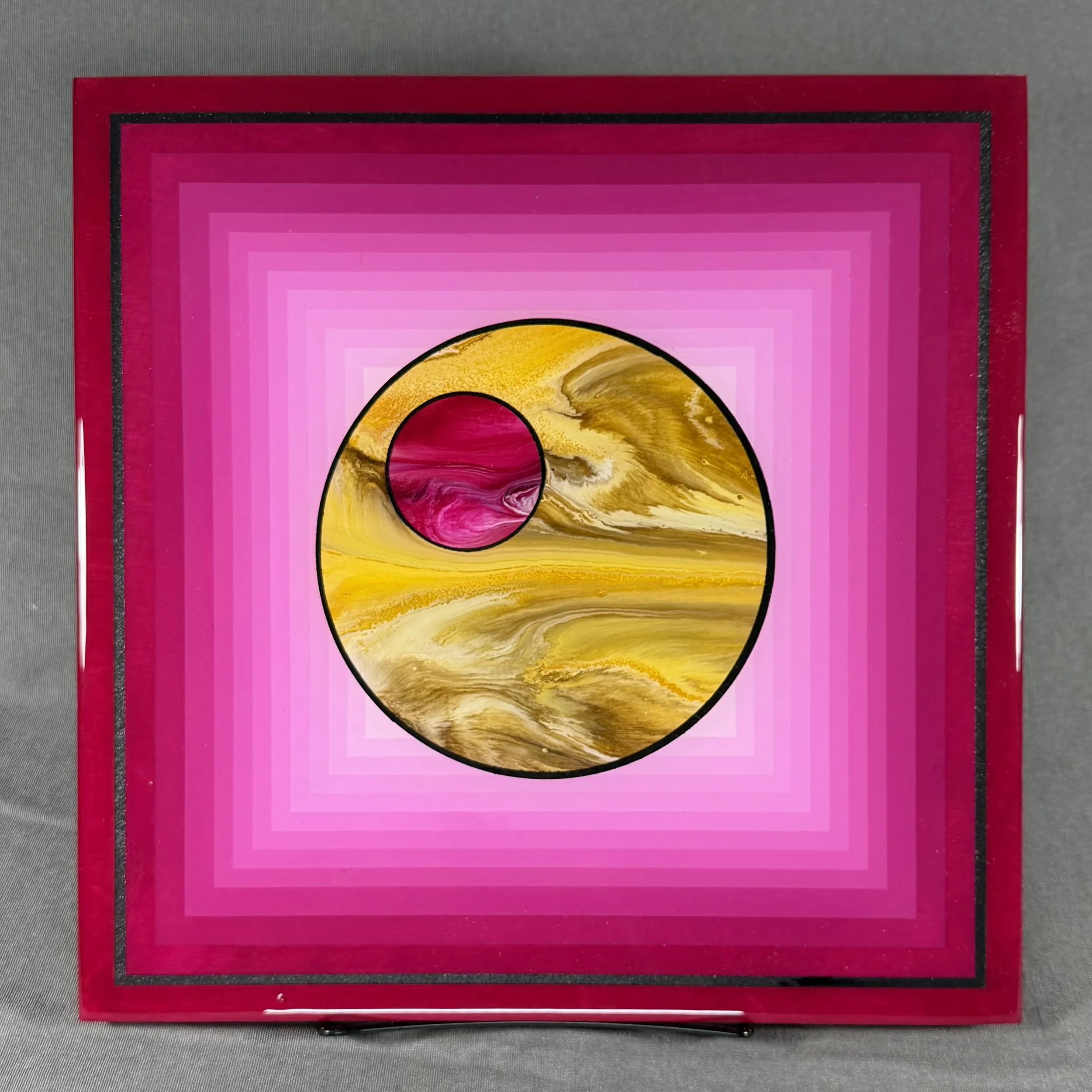 Colorful artwork with pink, yellow, and black in concentric circles, framed by red and pink borders, displayed on a gray surface.