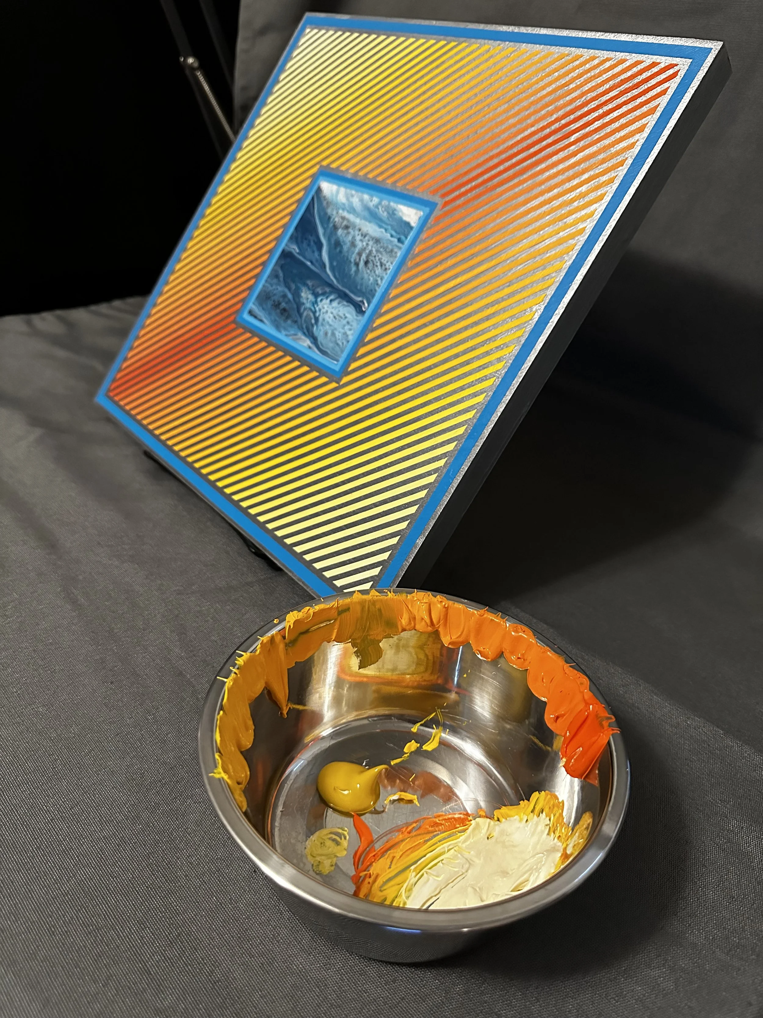 A metal mixing bowl containing yellow, orange, white, and red paint with some mixed paint at the bottom. In the background, a palette with a small square of swirling blue and white paint, and a colorful geometric artwork with diagonal stripes and a small rectangular section of ocean waves.