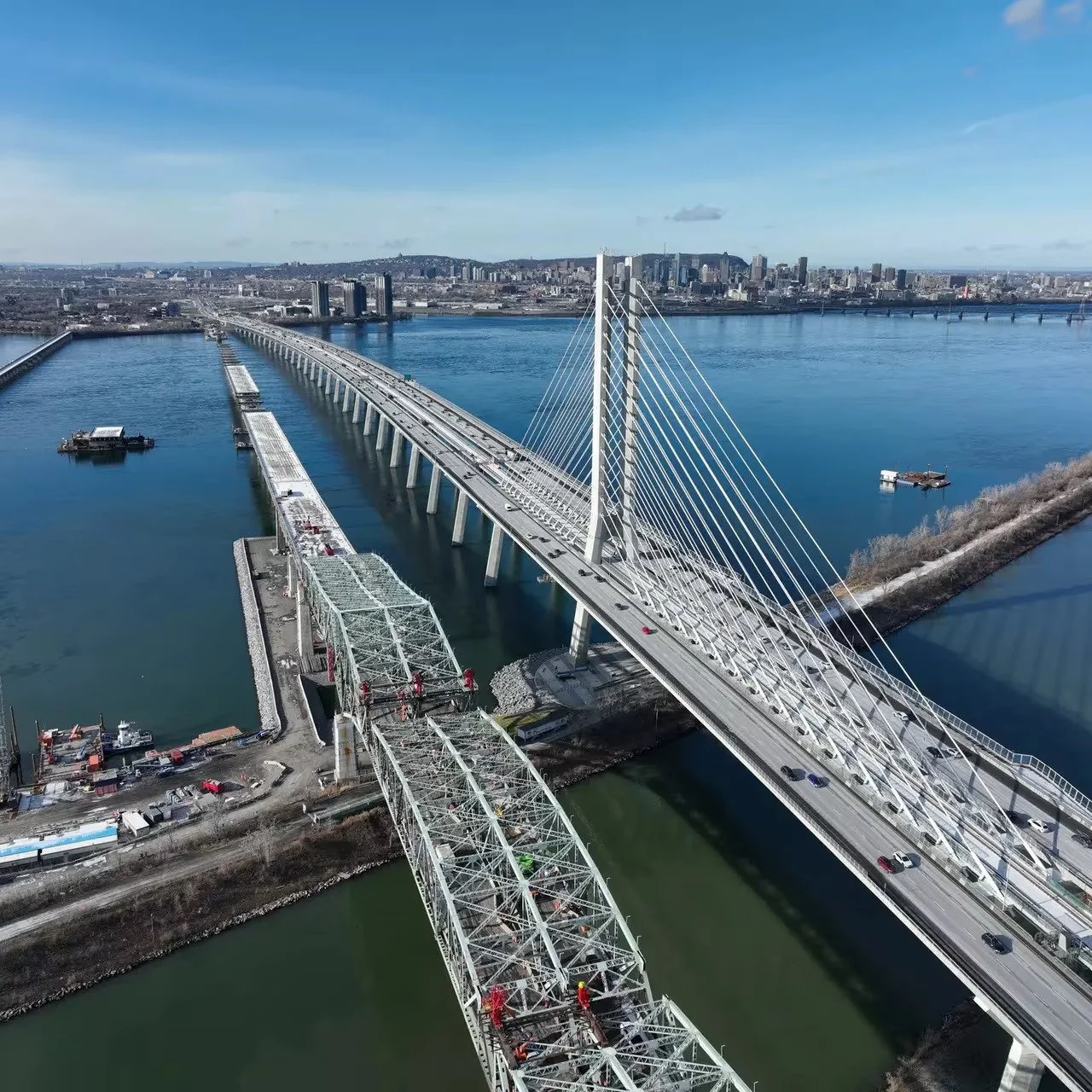 An aerial view of a modern cable-stayed bridge over a large river with a city skyline in the background. The bridge is under construction with visible cranes and construction equipment.