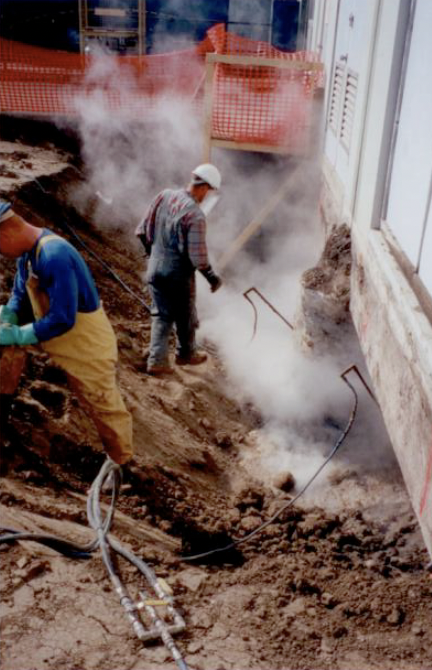 Construction workers are working inside a building, with dust and steam in the air, and some equipment and orange safety barriers around.