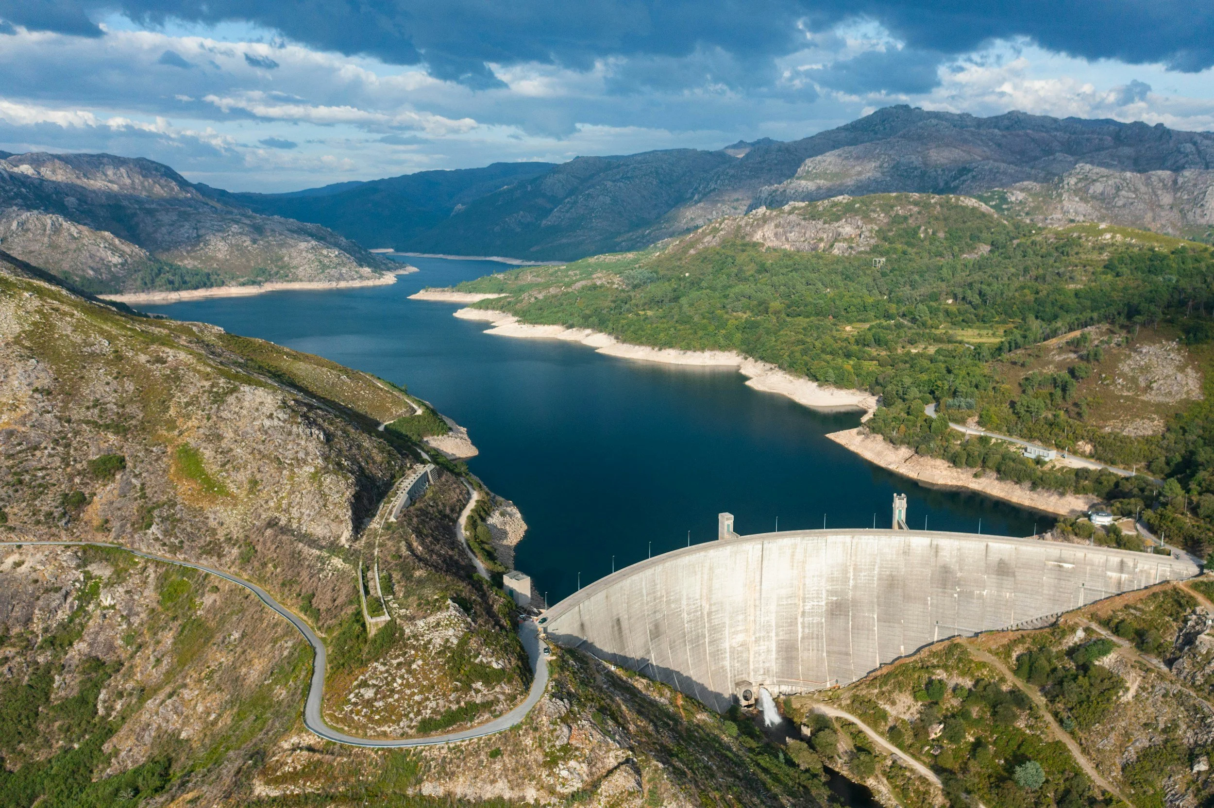 A large dam holding back a deep blue reservoir surrounded by green hills and mountains, with a cloudy sky overhead.
