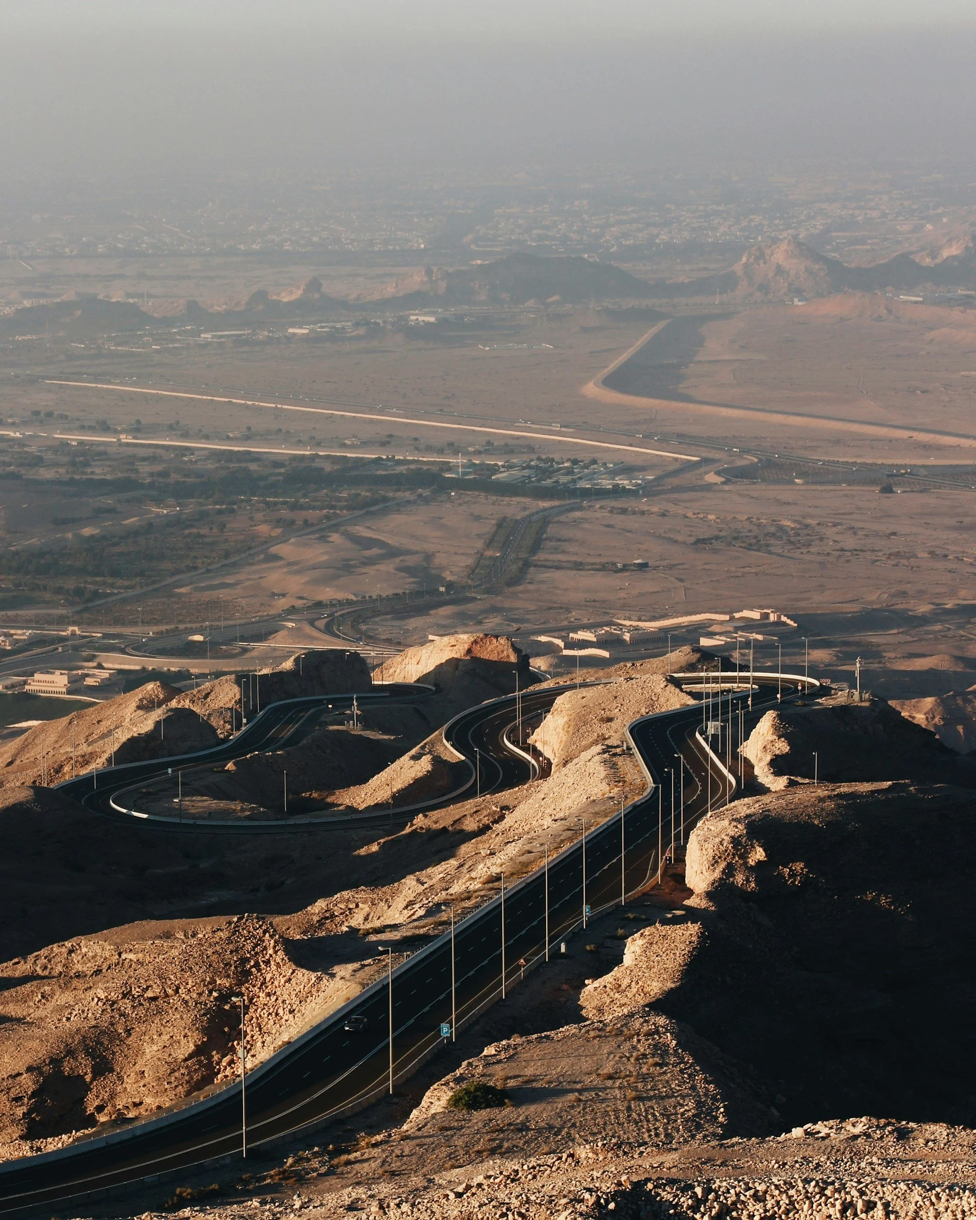 View of a winding road in a desert landscape with hills and mountains in the distance, under a hazy sky.