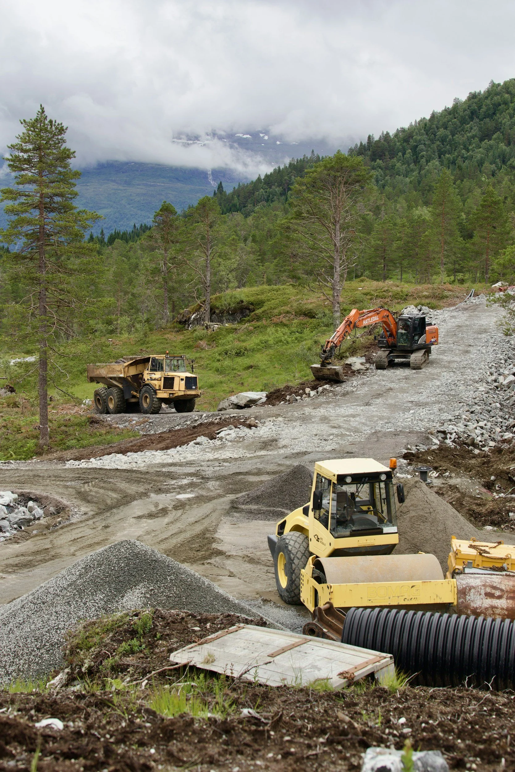 Construction site in a mountainous area with trees, gravel, and construction machinery, including a yellow bulldozer, an orange excavator, and a smaller vehicle, surrounded by greenery and misty mountains in the background.