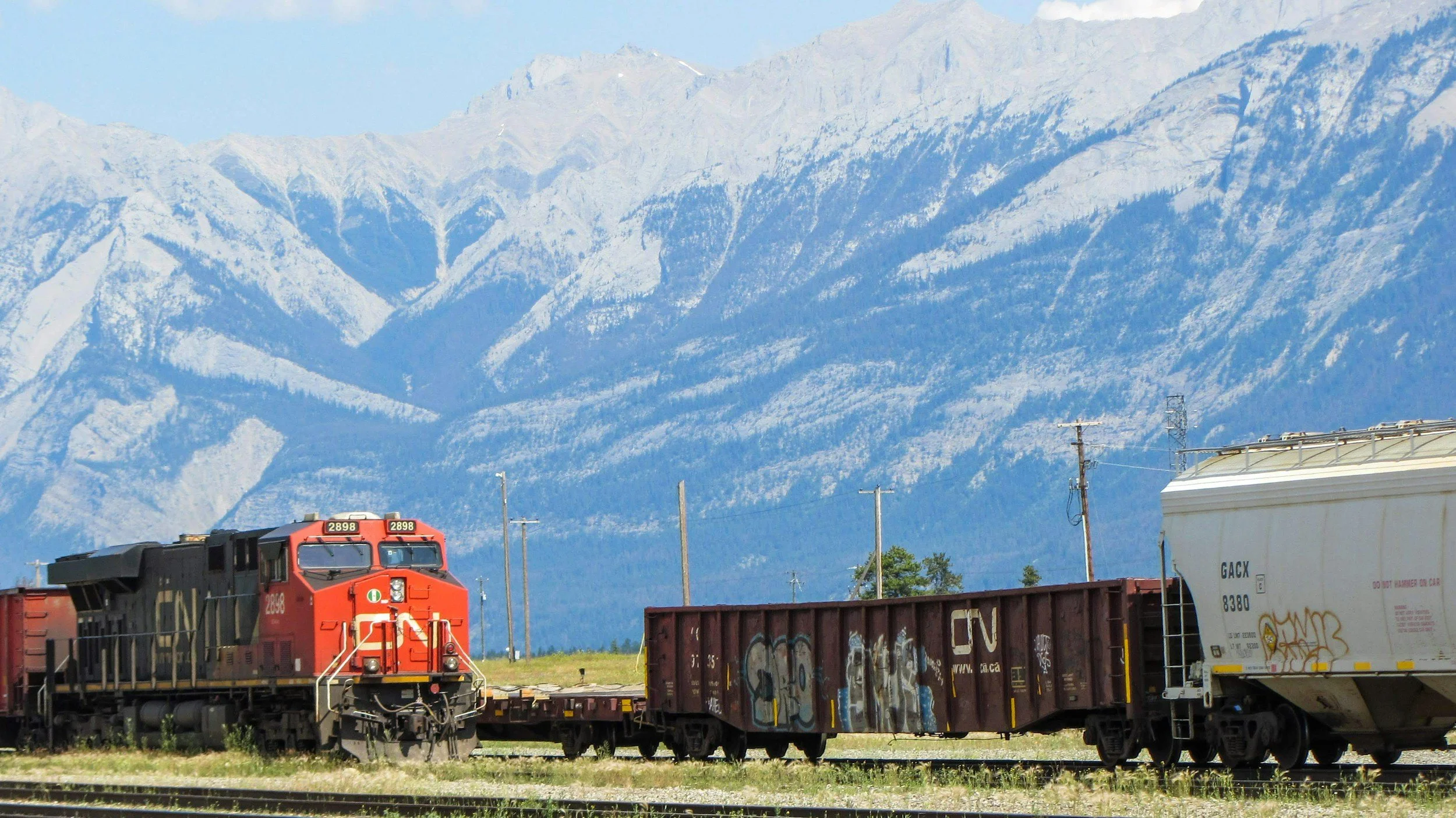 A red Canadian National freight train moving through a rural area with snowy mountain range in the background.