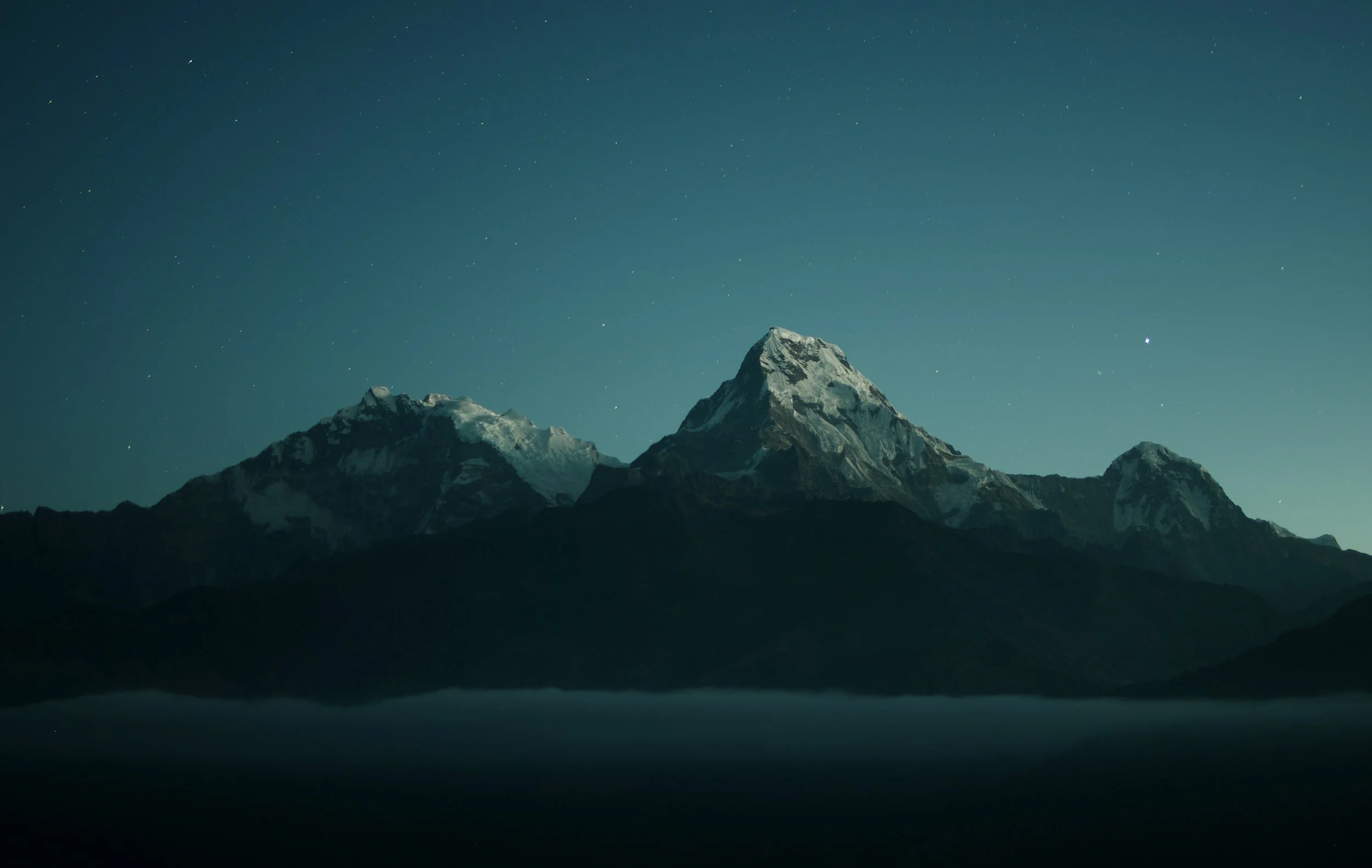 Nighttime view of snow-capped mountains with clear skies and stars above, reflecting on a body of water below.