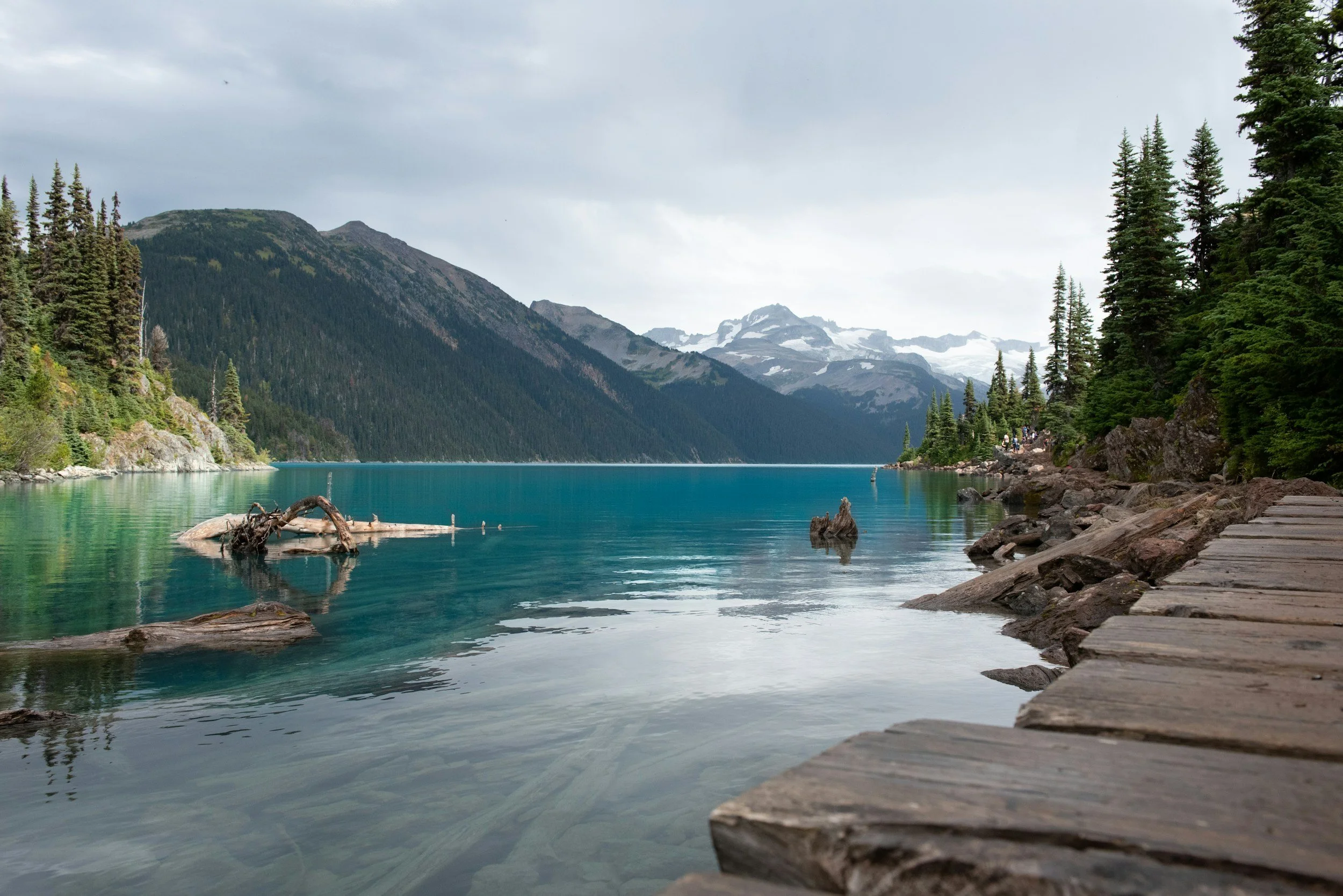 Scenic view of a mountain lake with clear turquoise water, surrounded by evergreen trees and snow-capped mountains in the background, with a cloudy sky overhead.