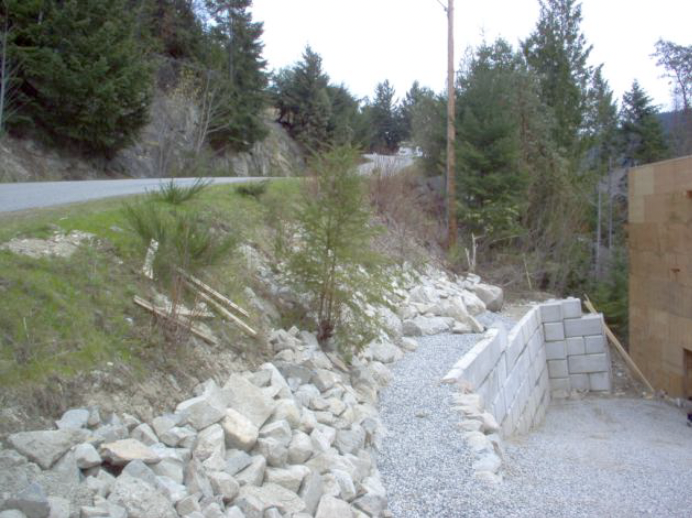 Construction site with a gravel footpath, stacked concrete blocks, rocks, and trees on a hillside.