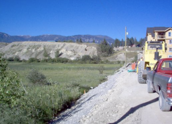 Construction vehicles and trucks on a gravel road beside green fields with mountains in the background.