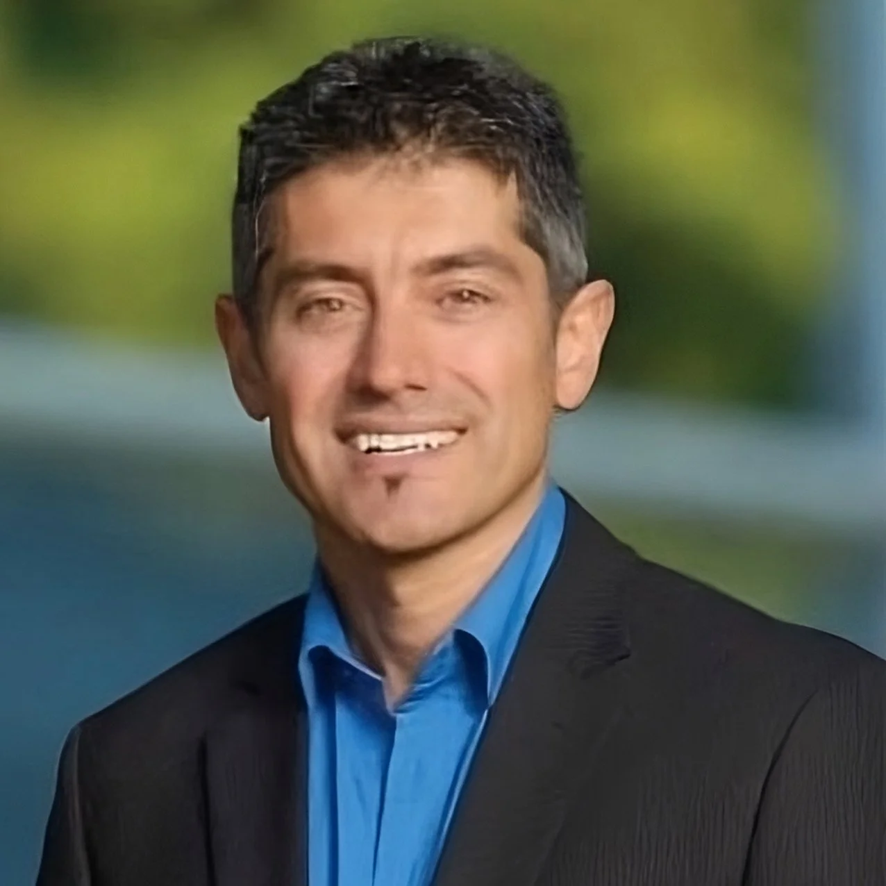 A smiling man with short dark hair, wearing a black suit jacket and a blue shirt, standing outdoors with a blurred background of green and blue.