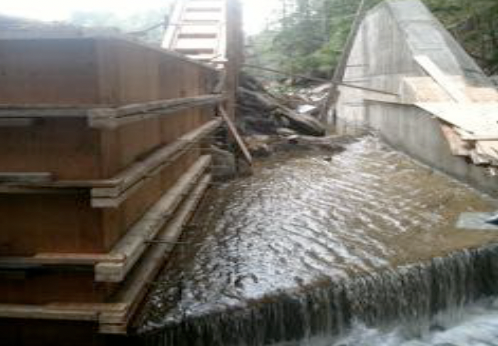 Flooded area with water flowing over a small dam or barrier, surrounded by wooden and concrete structures.