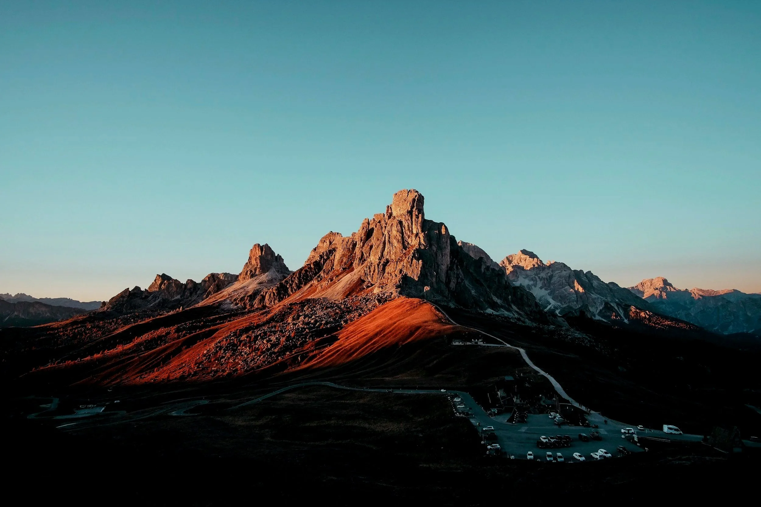 A rugged mountain range with sharp peaks illuminated by the setting or rising sun, with a parking lot and winding roads in the foreground at the base of the mountains, under a clear blue sky.