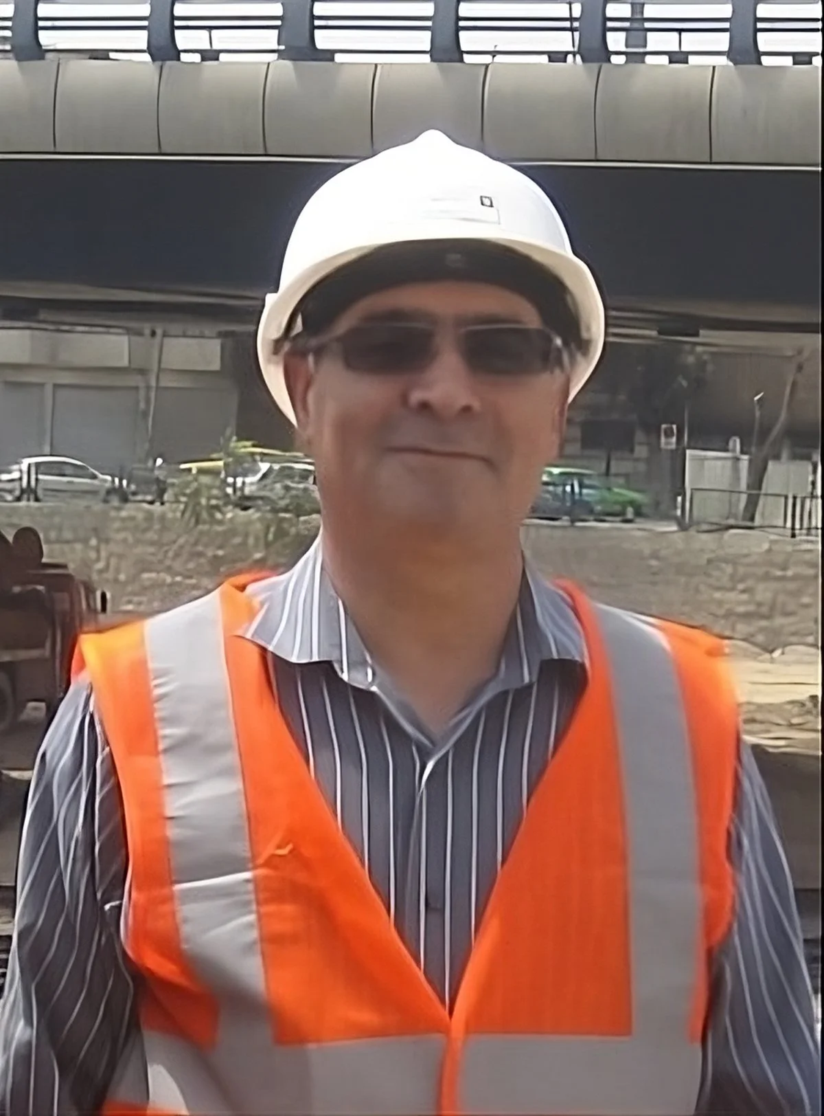 A man wearing a white safety helmet, dark sunglasses, and an orange safety vest with gray reflective stripes. He is outdoors at a construction site or industrial area.