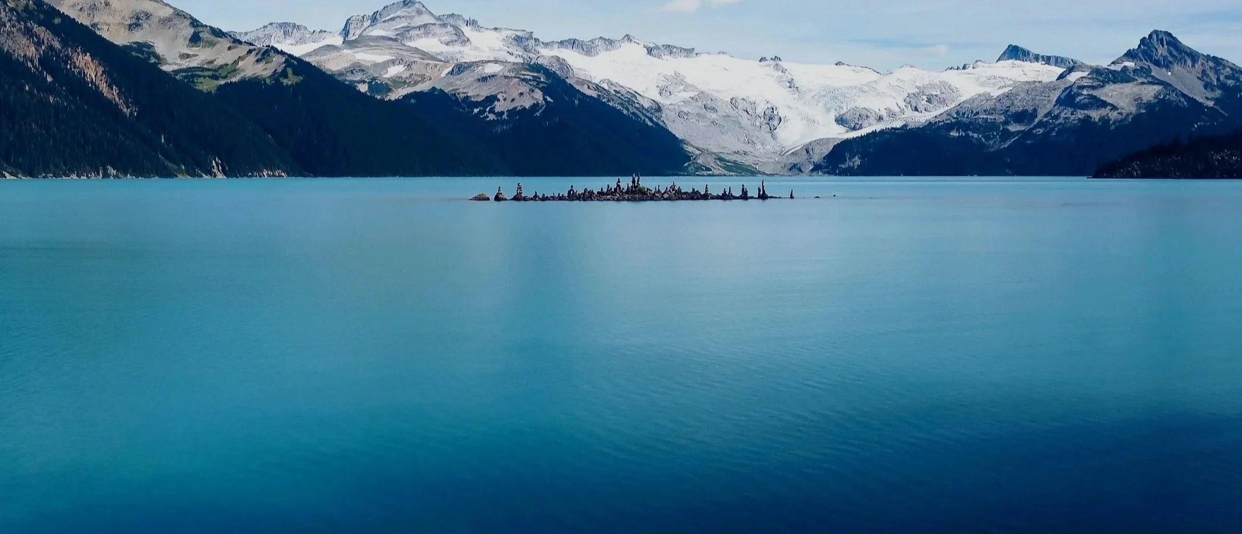 A serene lake with turquoise waters, surrounded by snow-capped mountains under a partly cloudy sky, with a small rocky island in the middle of the lake.