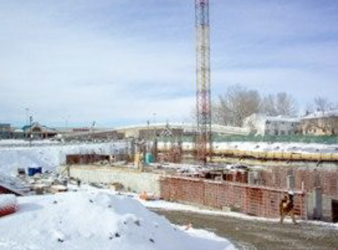 Snow-covered construction site with a tall communication tower and buildings in the background.