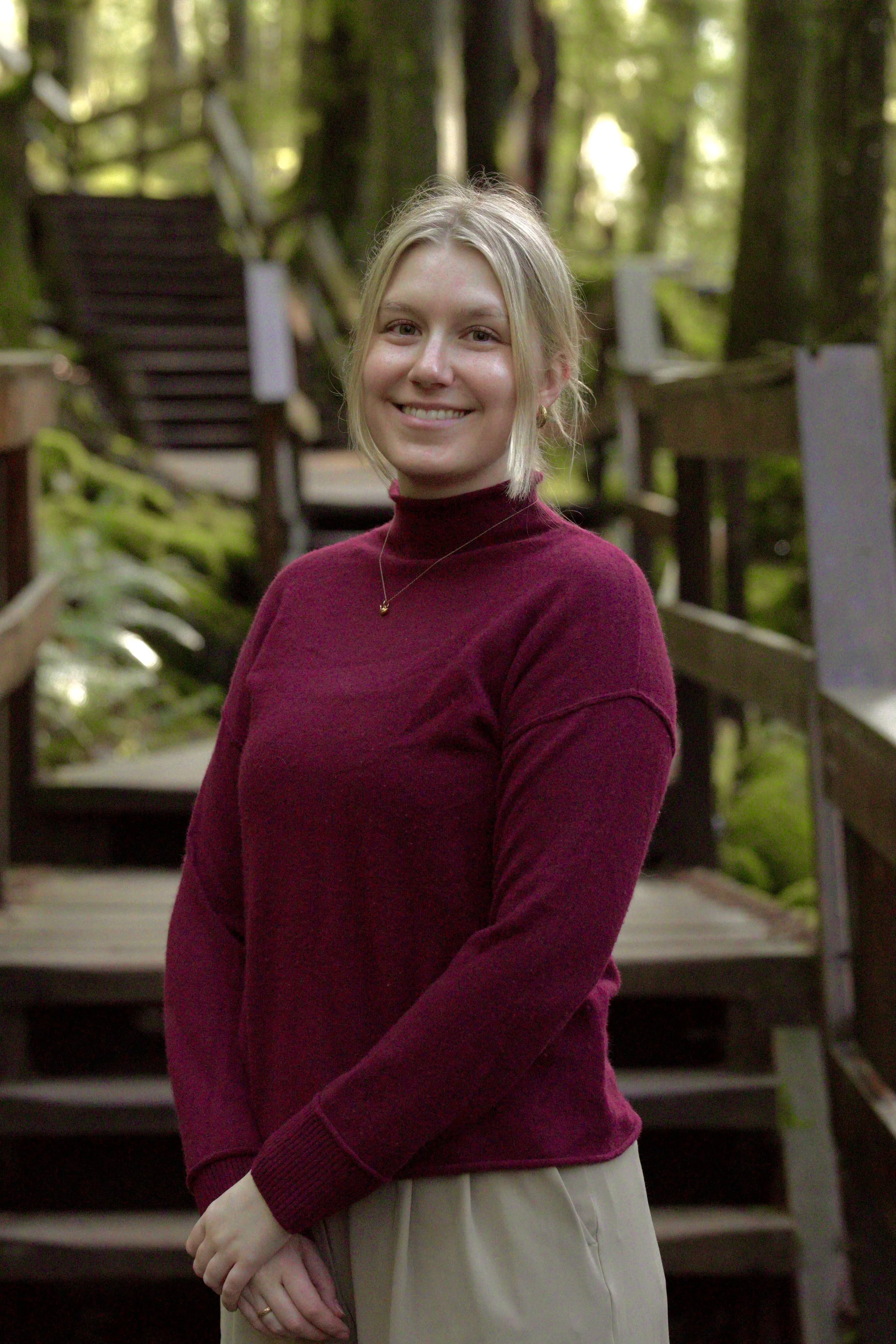 A smiling blonde woman wearing a maroon turtleneck sweater standing outdoors on a wooden staircase surrounded by green trees.