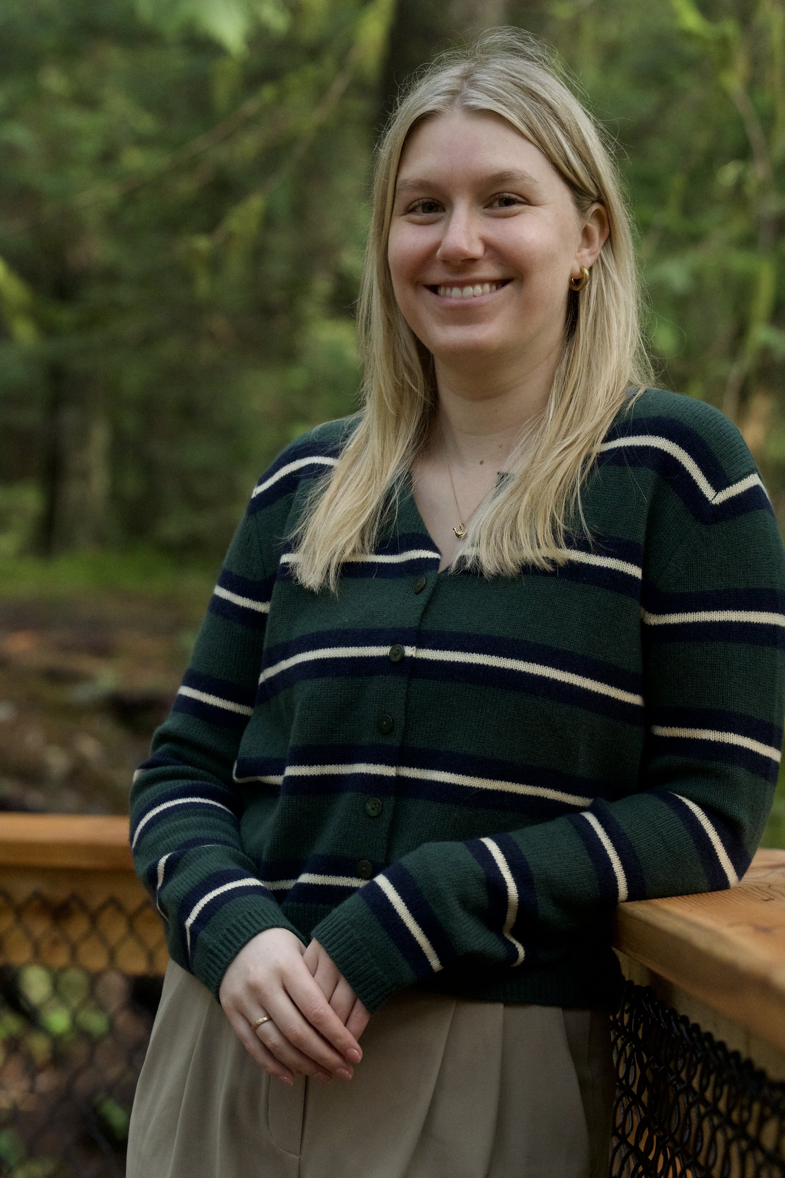 A woman with long blonde hair smiling outdoors, wearing a green and navy striped cardigan and a beige skirt, standing by a wooden railing in a park or garden setting with trees and greenery in the background.