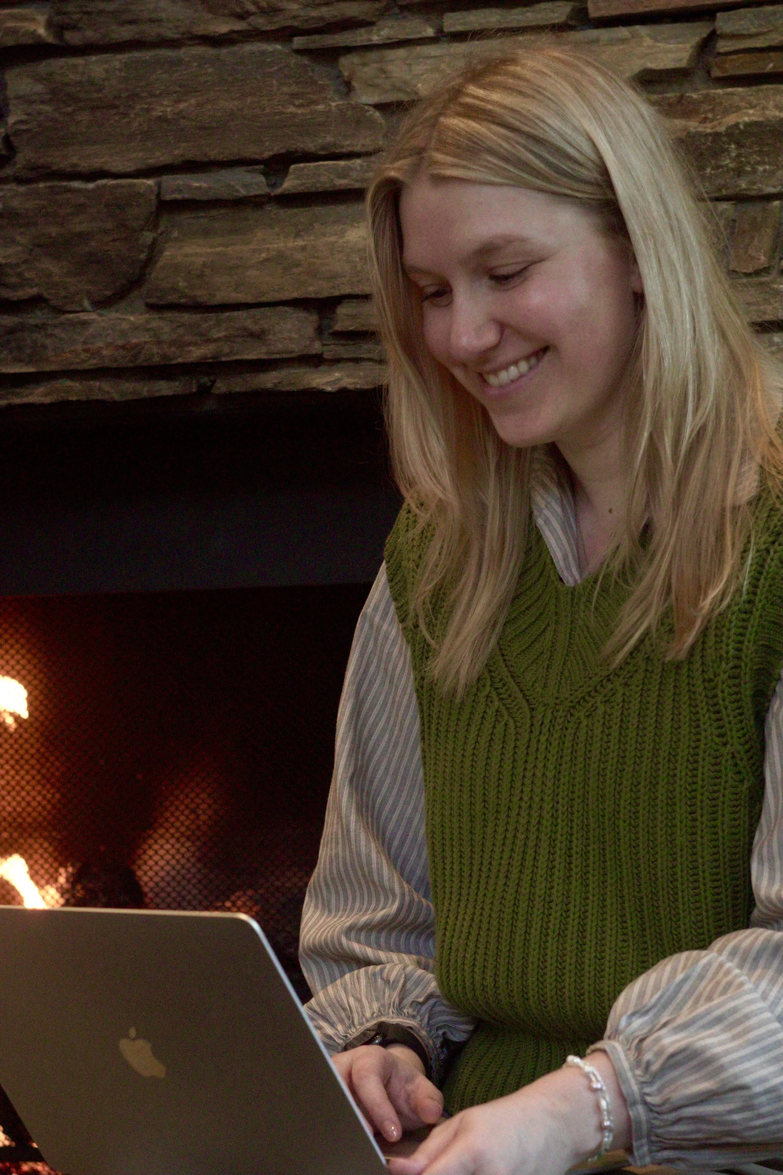 A woman with long blonde hair, wearing a green vest over a striped shirt, sitting in front of a fireplace with glowing embers, working on a laptop.