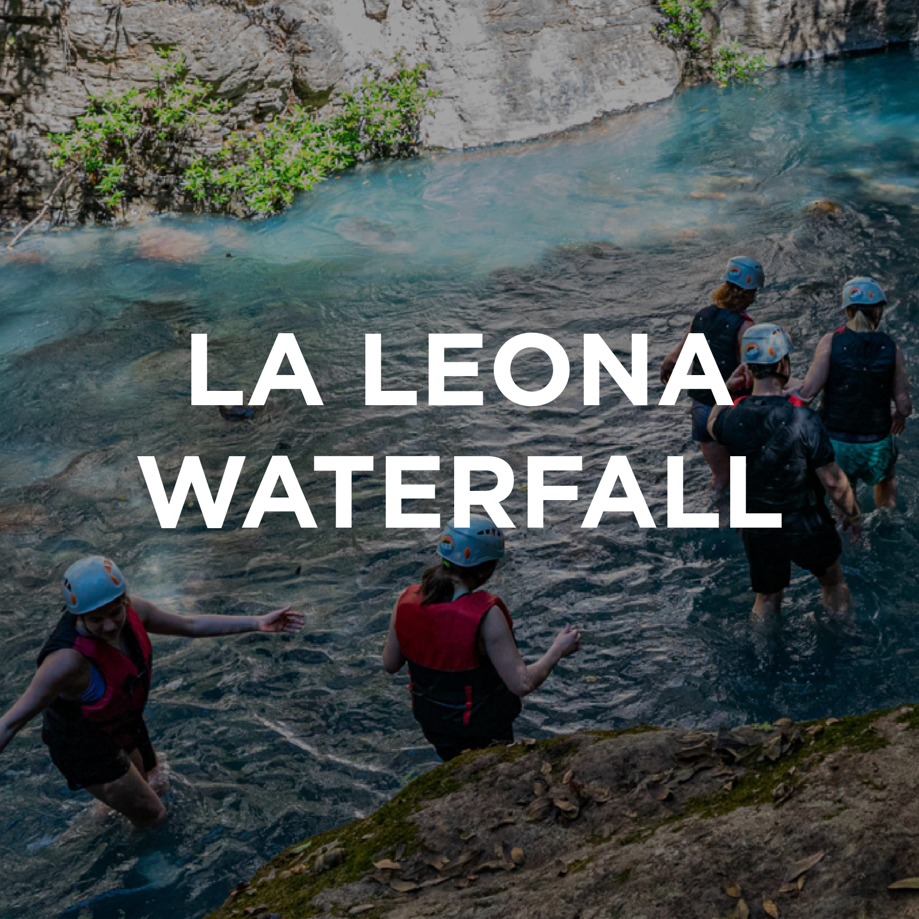 People in wetsuits and helmets wade through water near rocky and wooded riverbank, preparing for river activity at La Leona Waterfall.