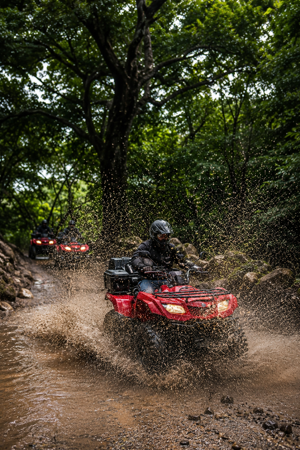 A person riding a red ATV through a muddy trail in a forest, splashing water as they drive, with other riders visible in the background.