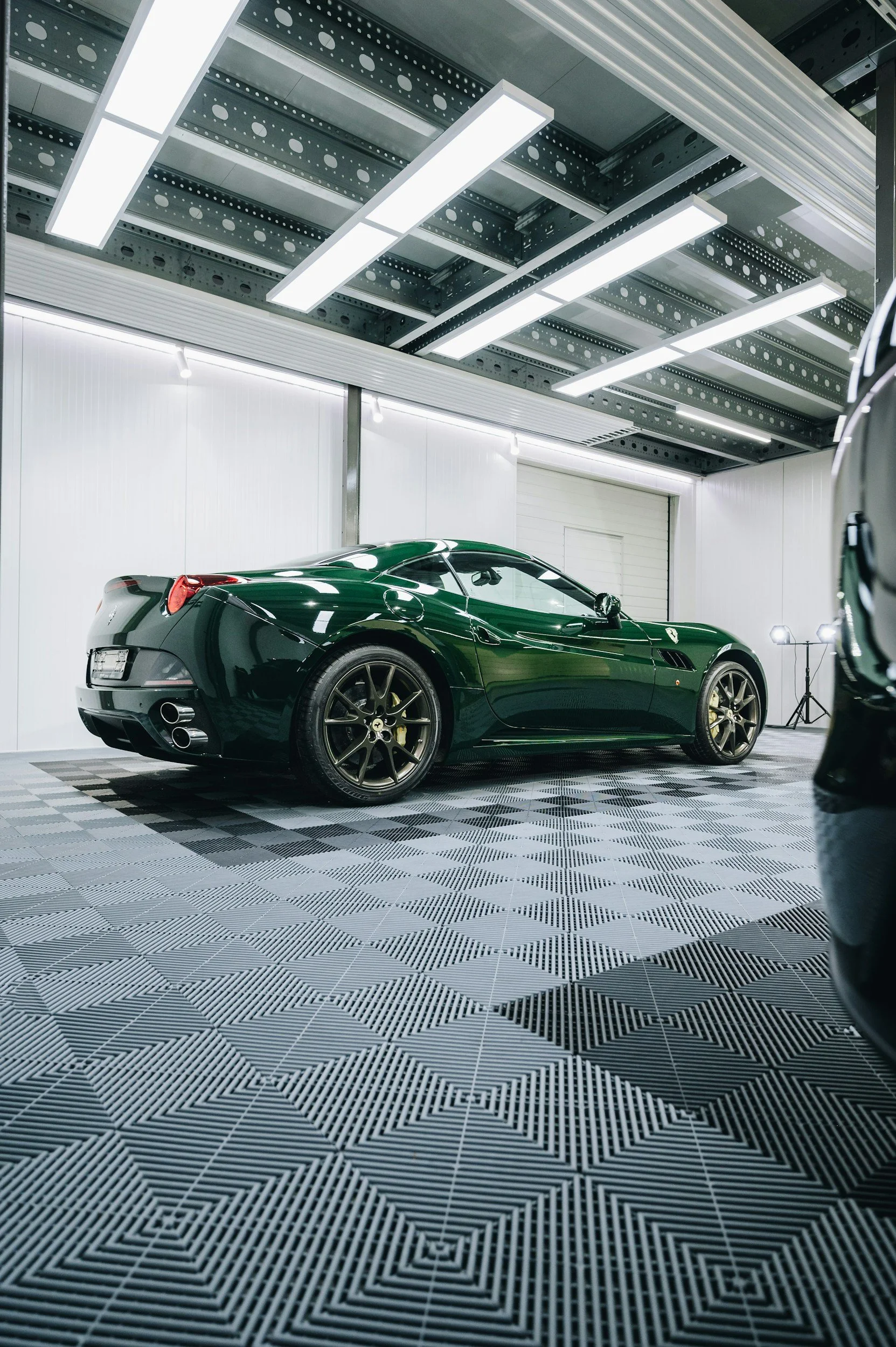 A black sports car parked inside a well-lit garage with a patterned rubber floor and a metallic ceiling.