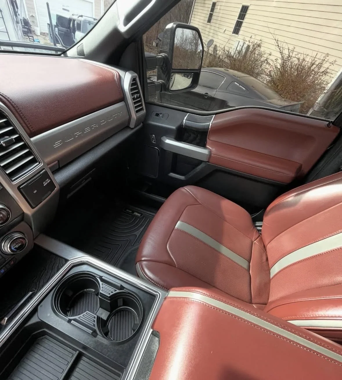 Inside the cab of a vehicle, showing a red leather seat, dashboard area with steering wheel, and side mirrors. Outside the window, a building and some bushes are visible.